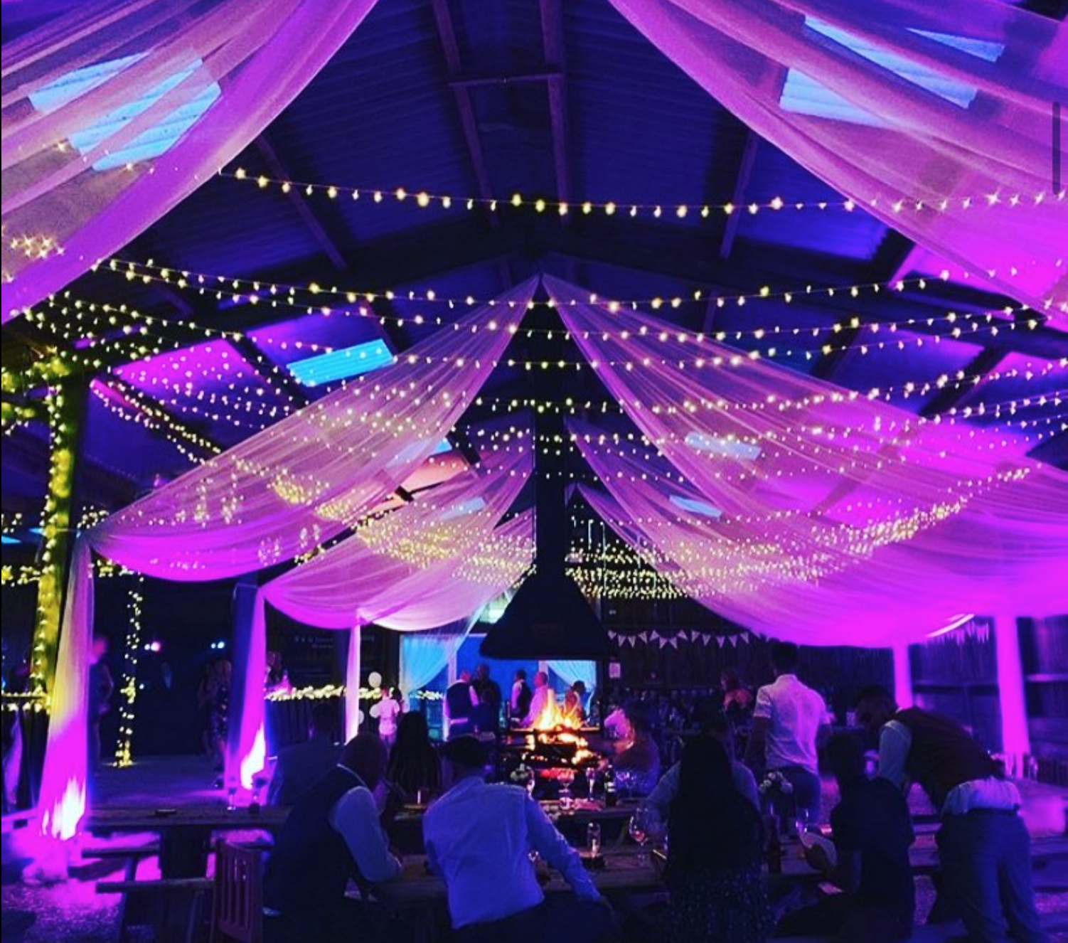 People gathered under a purple and pink draped barn decorated with string lights at a nighttime event, with a fire pit in the center.