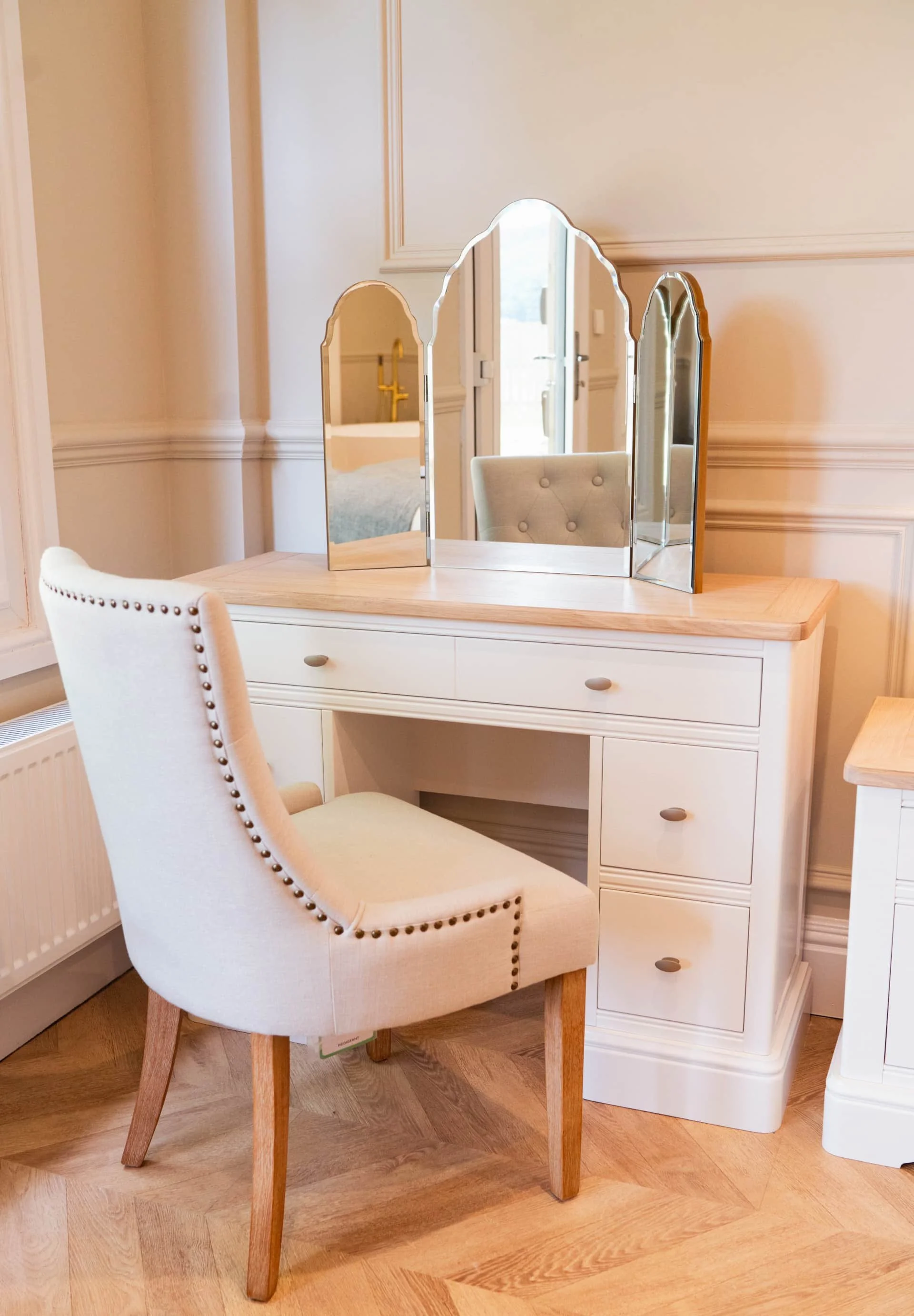 A beige upholstered chair with wooden legs placed in front of a white vanity table with a wooden top. The vanity has three drawers with round handles and a three-panel decorative mirror on top. The background features cream-colored walls with decorative molding.