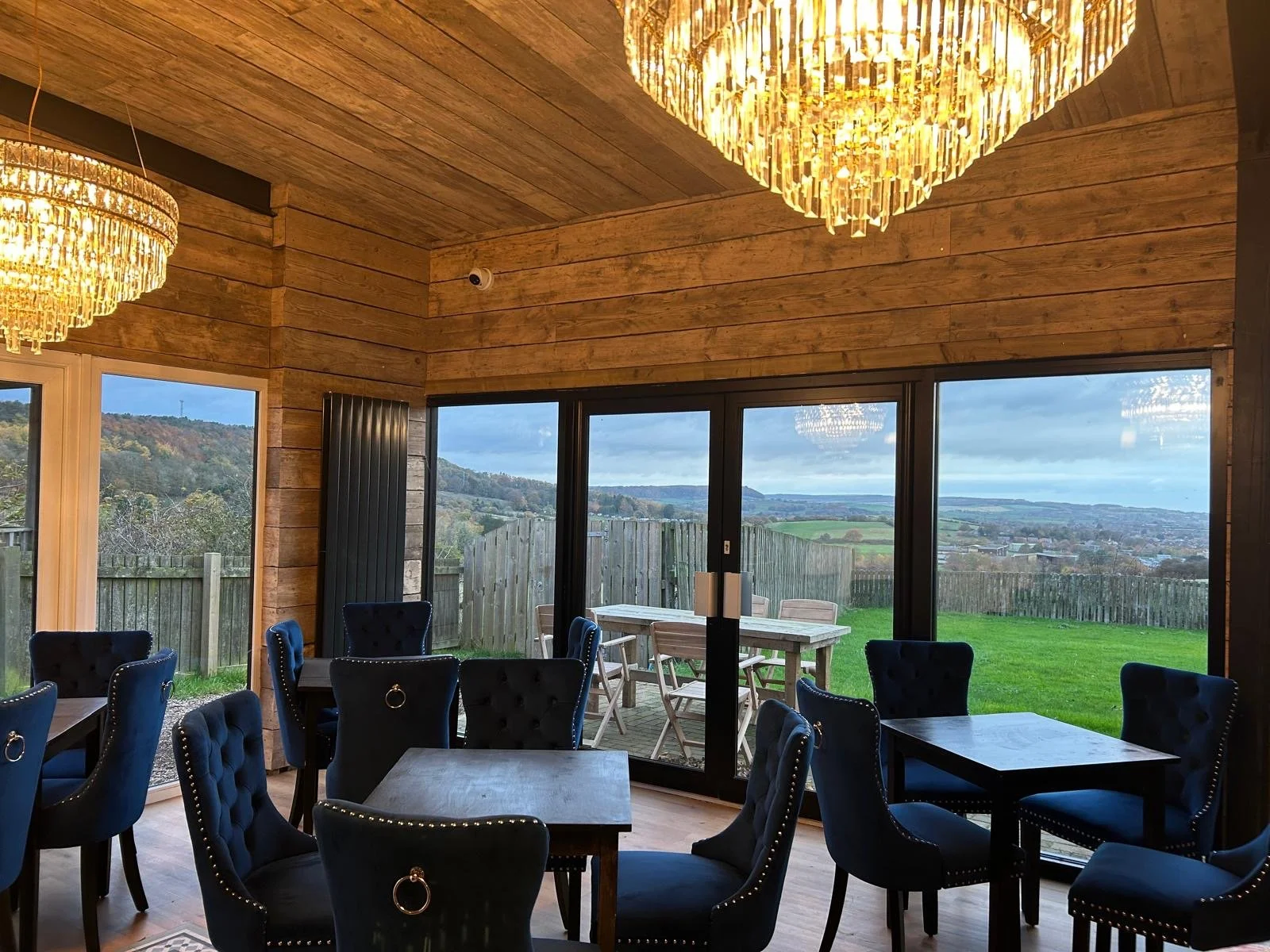 Interior of Function Room chairs around dark wooden tables, wood-paneled walls, large sliding glass doors opening to a grassy area with a wooden fence and scenic hills in the distance, and two large gold chandeliers hanging from a wooden ceiling.