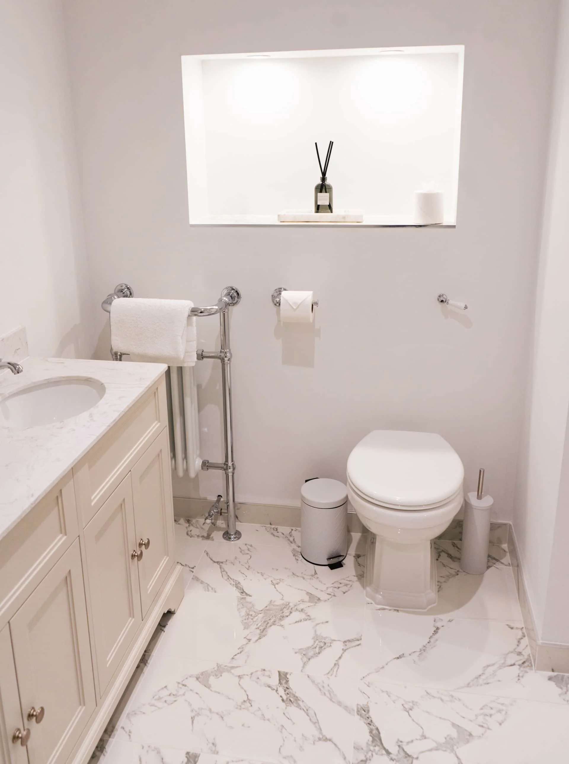A minimalist white bathroom with marble floor, a white vanity with a marble countertop, a toilet, a small white trash bin, a toilet brush, a towel on a chrome rack, and a recessed shelf with decorative items and a candle.