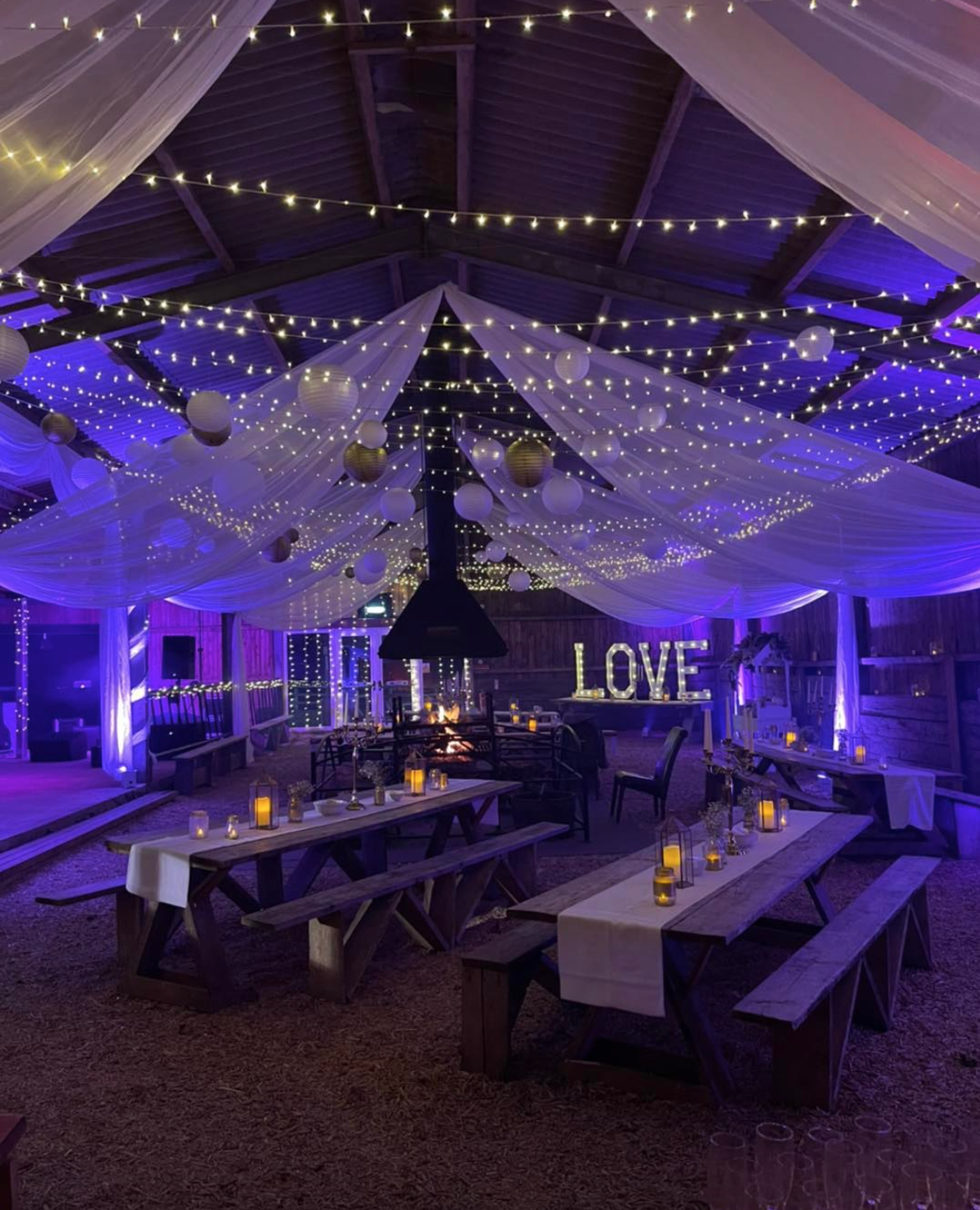 Decorated barn interior with string lights, hanging paper lanterns, draped fabric, candles, and a large illuminated "LOVE" sign, set for a celebration or wedding reception.