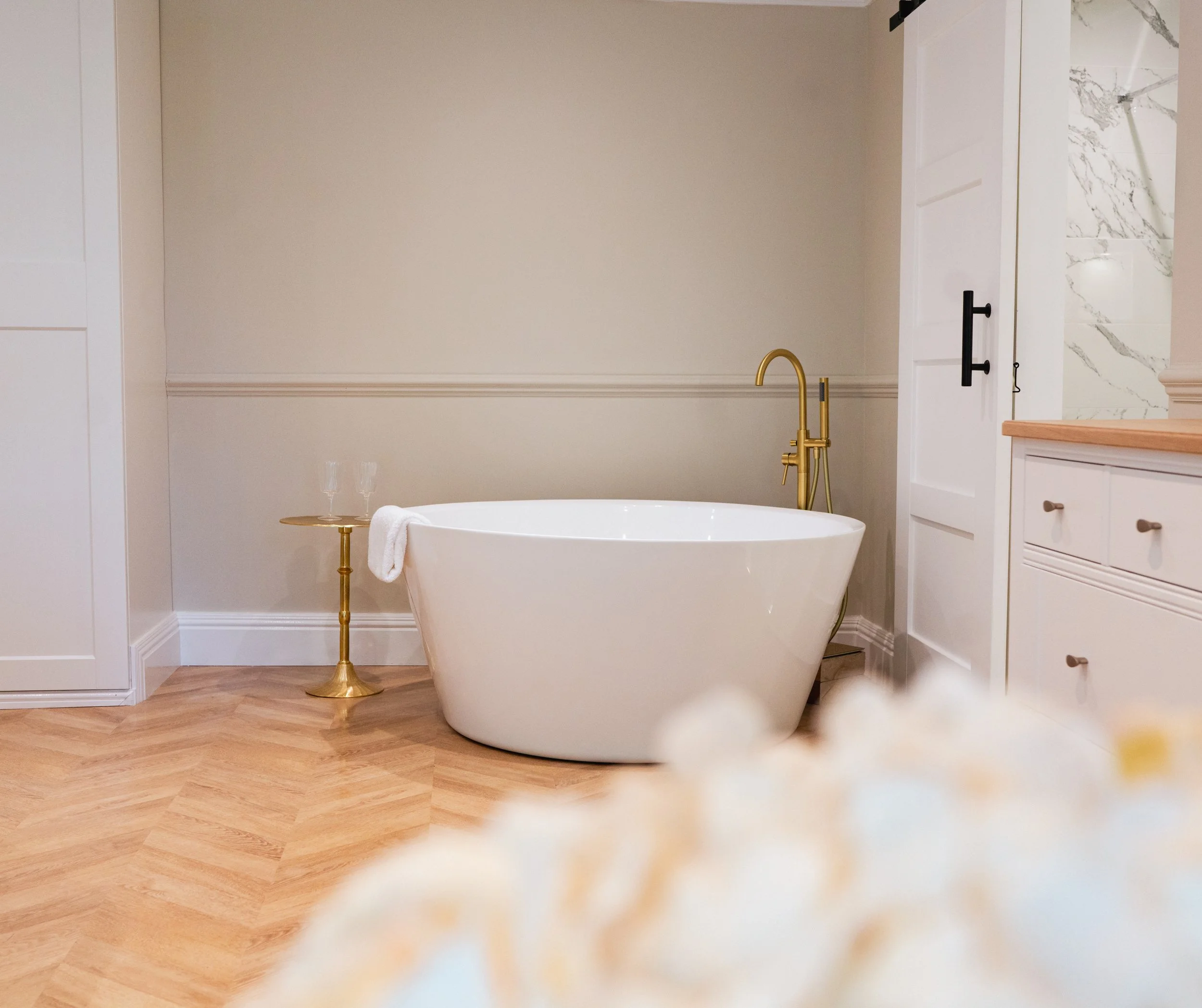 Modern bathroom with a freestanding white bathtub, gold fixtures, a small gold side table with two champagne glasses, and a wood floor.