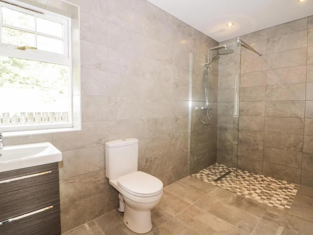 Modern bathroom with beige tiled walls and floor, a white toilet, a small vanity with a white sink, a window with a view of greenery, and a walk-in shower with a glass partition and a rain showerhead.