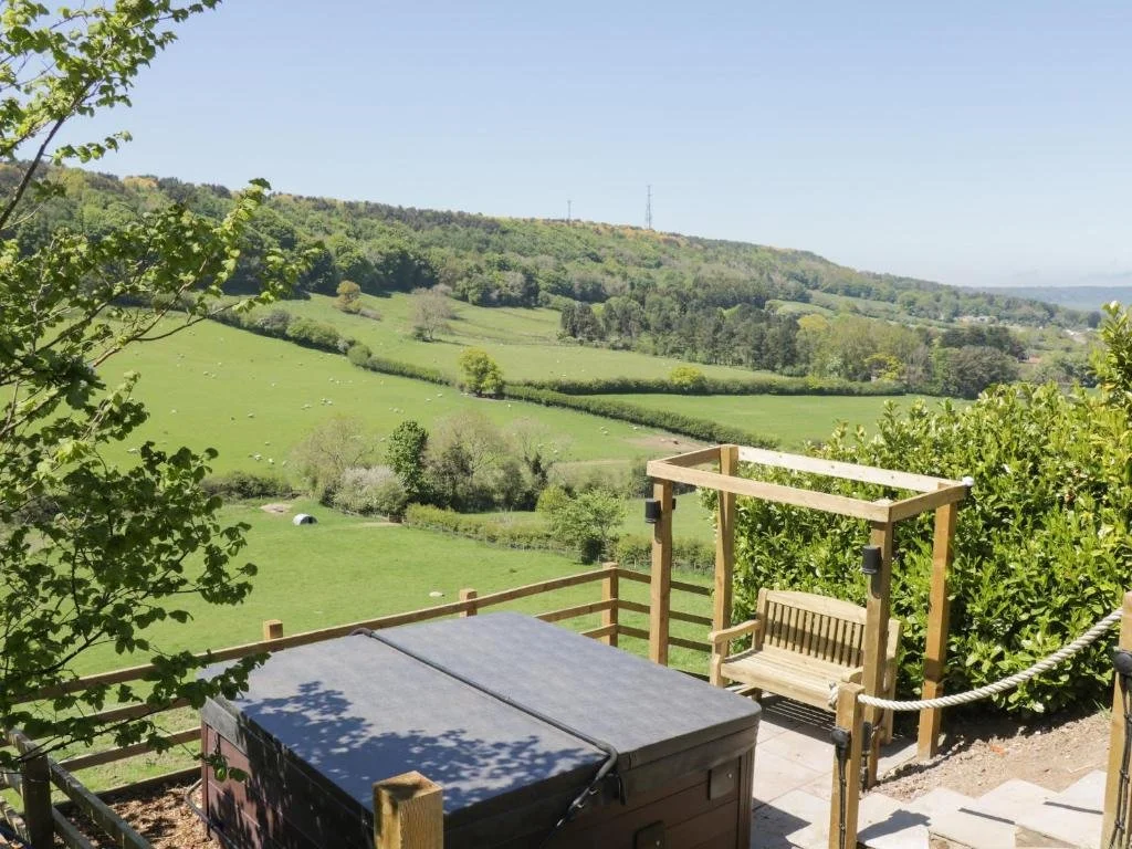 A terrace overlooking green rolling hills with trees, bushes, and a few distant structures under a clear blue sky.
