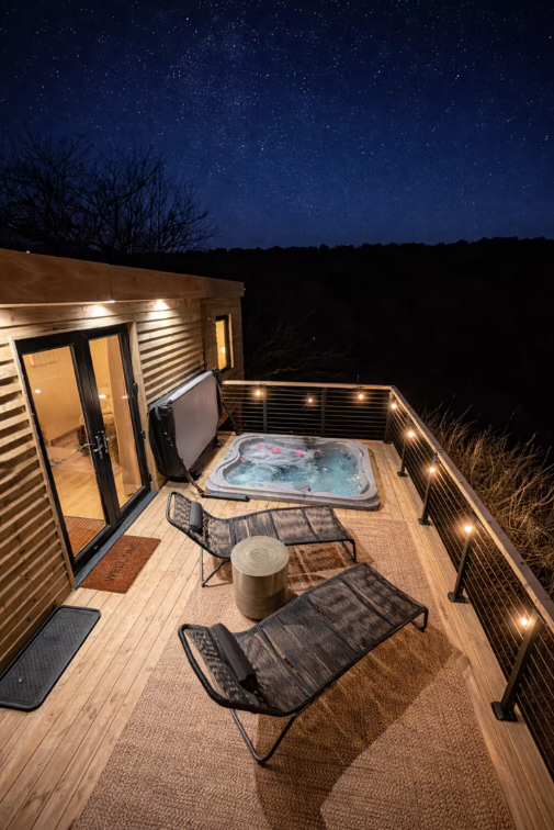 Nighttime view of a wooden balcony with two lounge chairs, a small round table, a hot tub, and string lights, with a starry sky in the background.