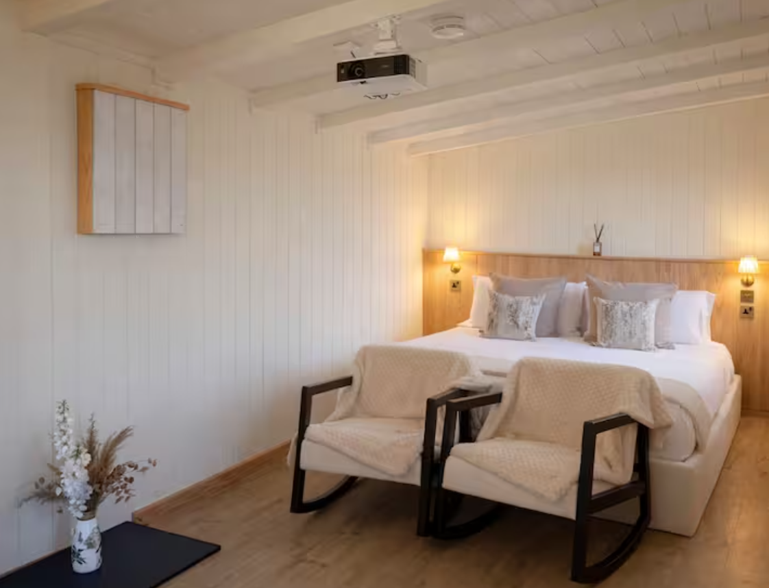 A bedroom with a white bed, pillows, and wooden headboard, two wall-mounted lamps, two upholstered chairs, a small black table with a white floral vase, and a wall-mounted speaker or art piece.