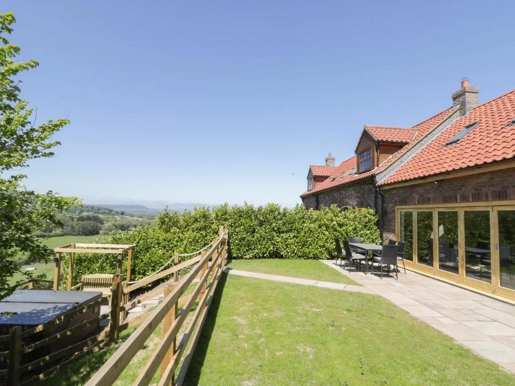 Backyard patio with outdoor dining table, chairs, and a grassy lawn, overlooking scenic countryside with green hills under a clear blue sky.