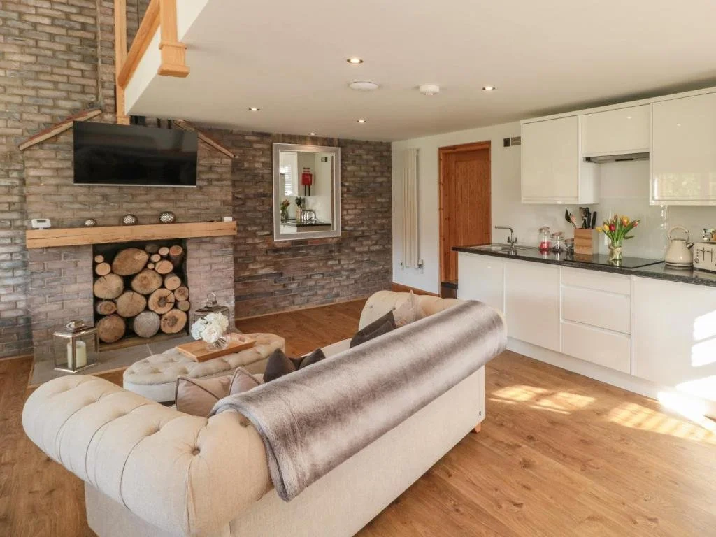 Living room with wood-lined fireplace, flat-screen TV, beige tufted sofa, and open kitchen with white cabinets, wooden accents, and hardwood floors.