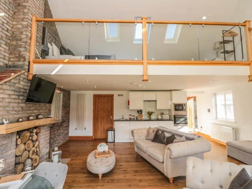 Open-plan living room with brick wall, wood accents, and a loft area with glass railing. Features a white sofa with dark pillows, a cozy round ottoman, and a fireplace with stacked logs. Kitchen with white cabinets and Stainless Steel appliances.