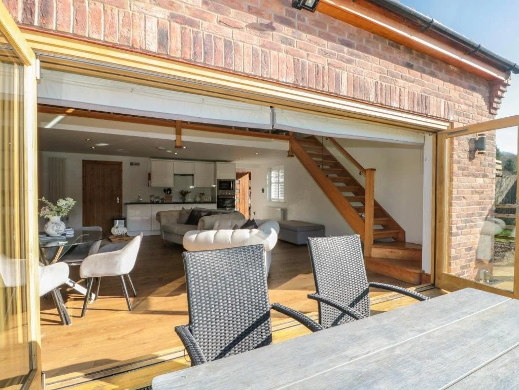Open sliding glass doors reveal a living space with a kitchen, dining, and seating area, wooden stairs, and natural sunlight.