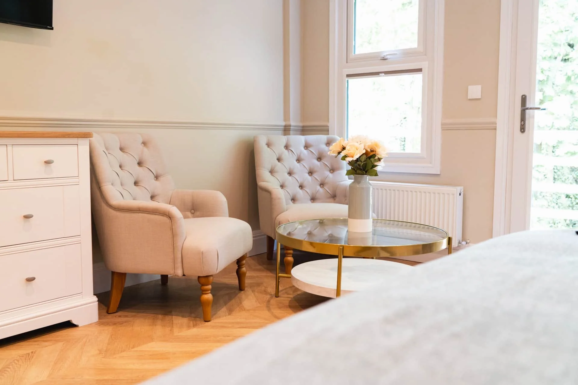Living room with two beige tufted armchairs, a glass-top coffee table with a gold frame, and a white vase with pink flowers, next to a window and a radiator.