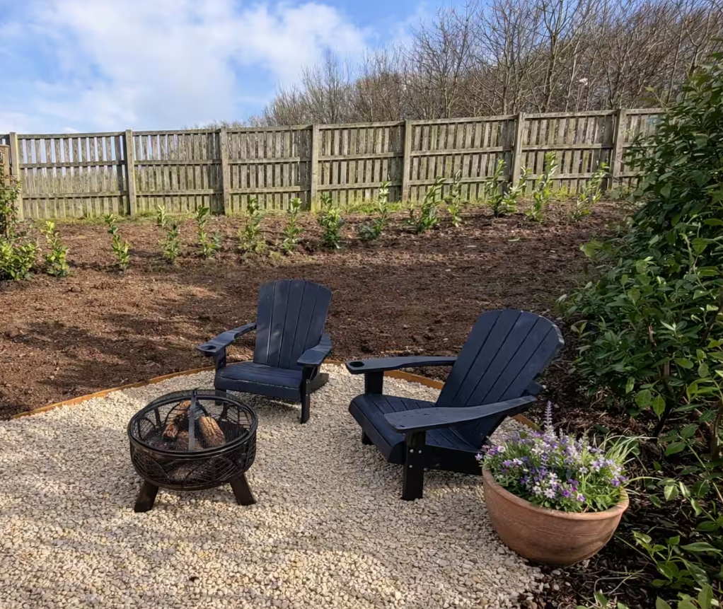 An outdoor seating area with two black Adirondack chairs and a small fire pit on a gravel surface, surrounded by a garden with a wooden fence, young plants, trees, and a potted flowering plant.