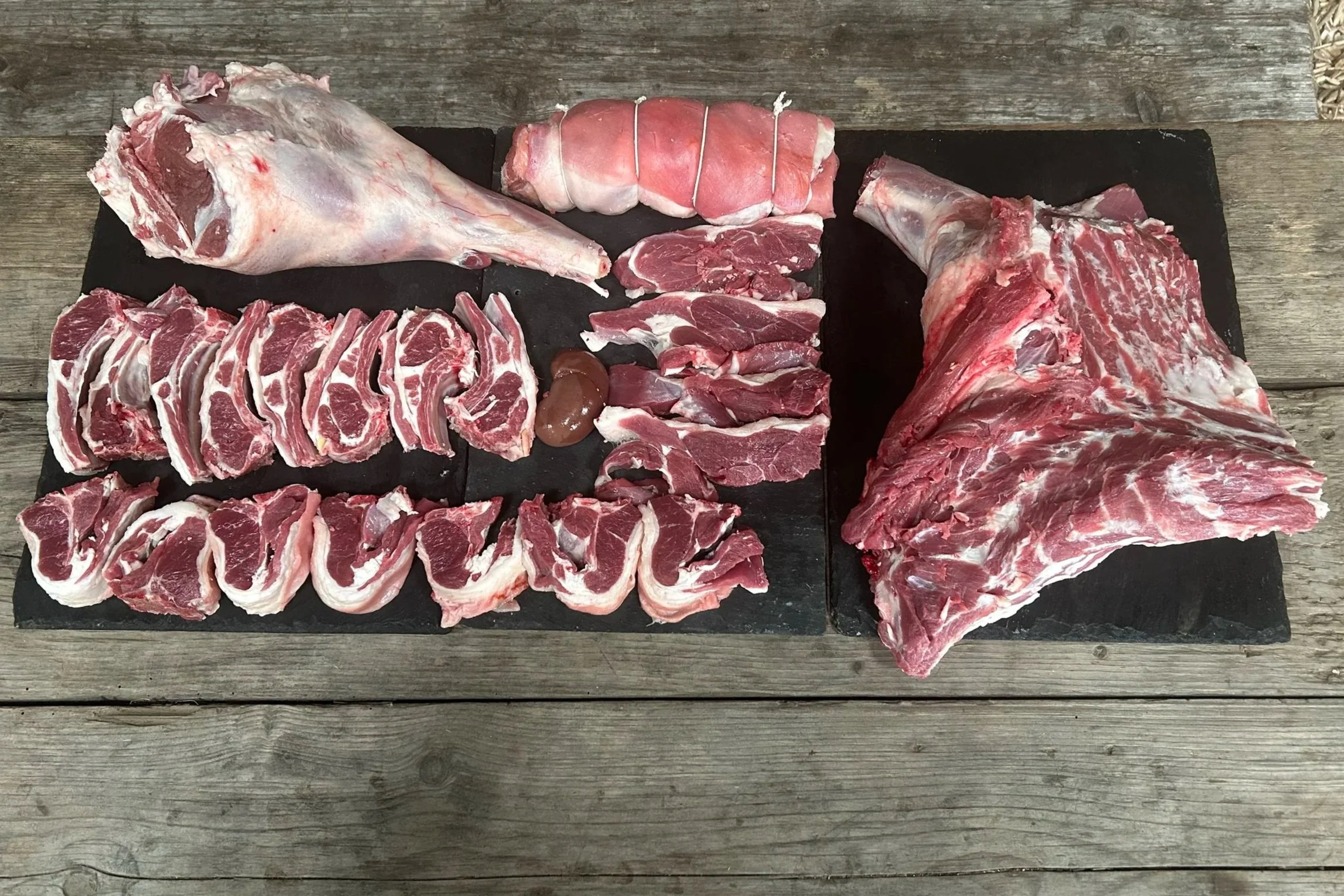 Various cuts of raw red meat, including ribs, steak, and pork shoulder, arranged on two black slabs on a wooden surface.