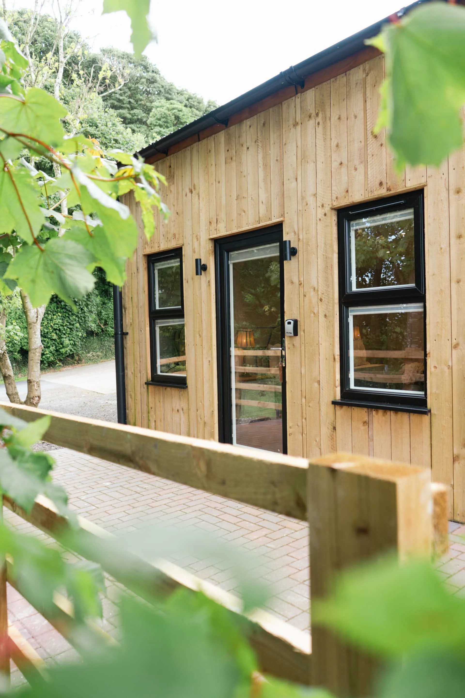 A wooden panelled building with black-framed windows and a glass door, surrounded by greenery and a wooden fence.