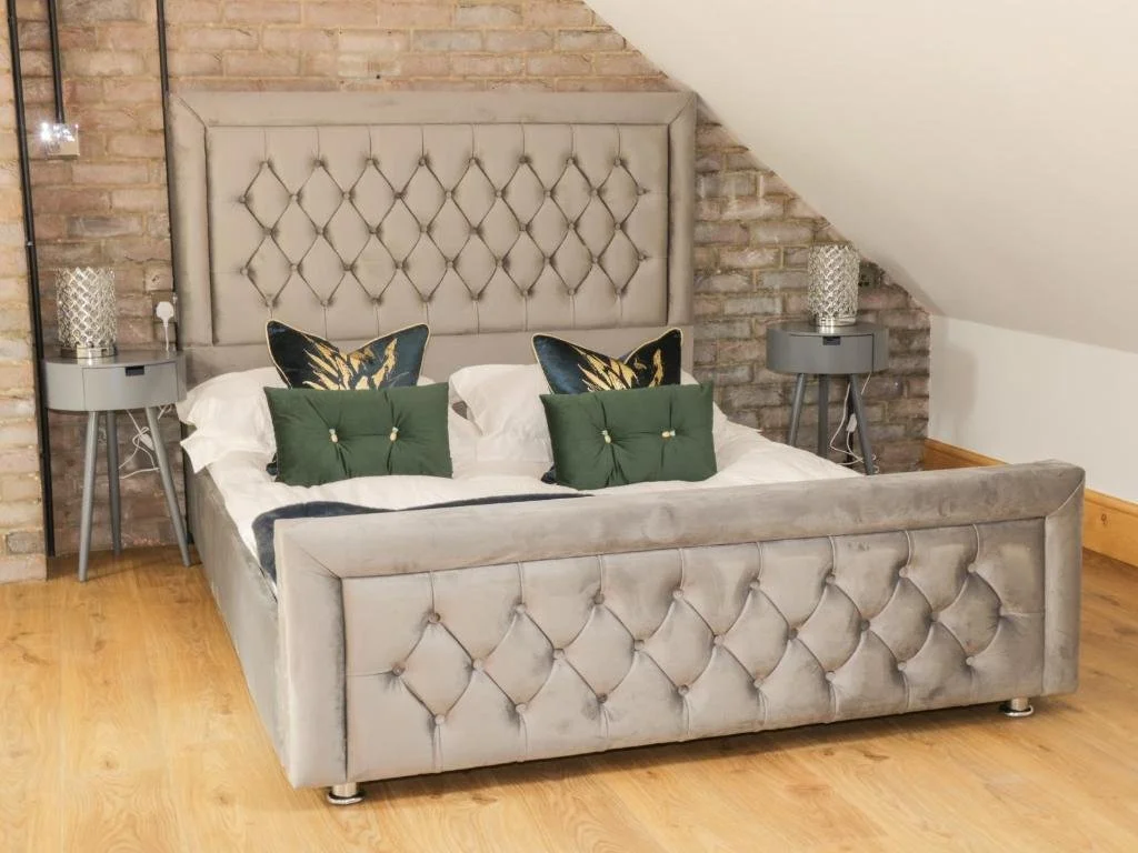 A bedroom with a large upholstered bed featuring a tufted headboard and footboard, decorated with white, green, and patterned pillows. The room has exposed brick walls and wooden flooring, with two gray nightstands on either side of the bed, each holding a decorative lamp.