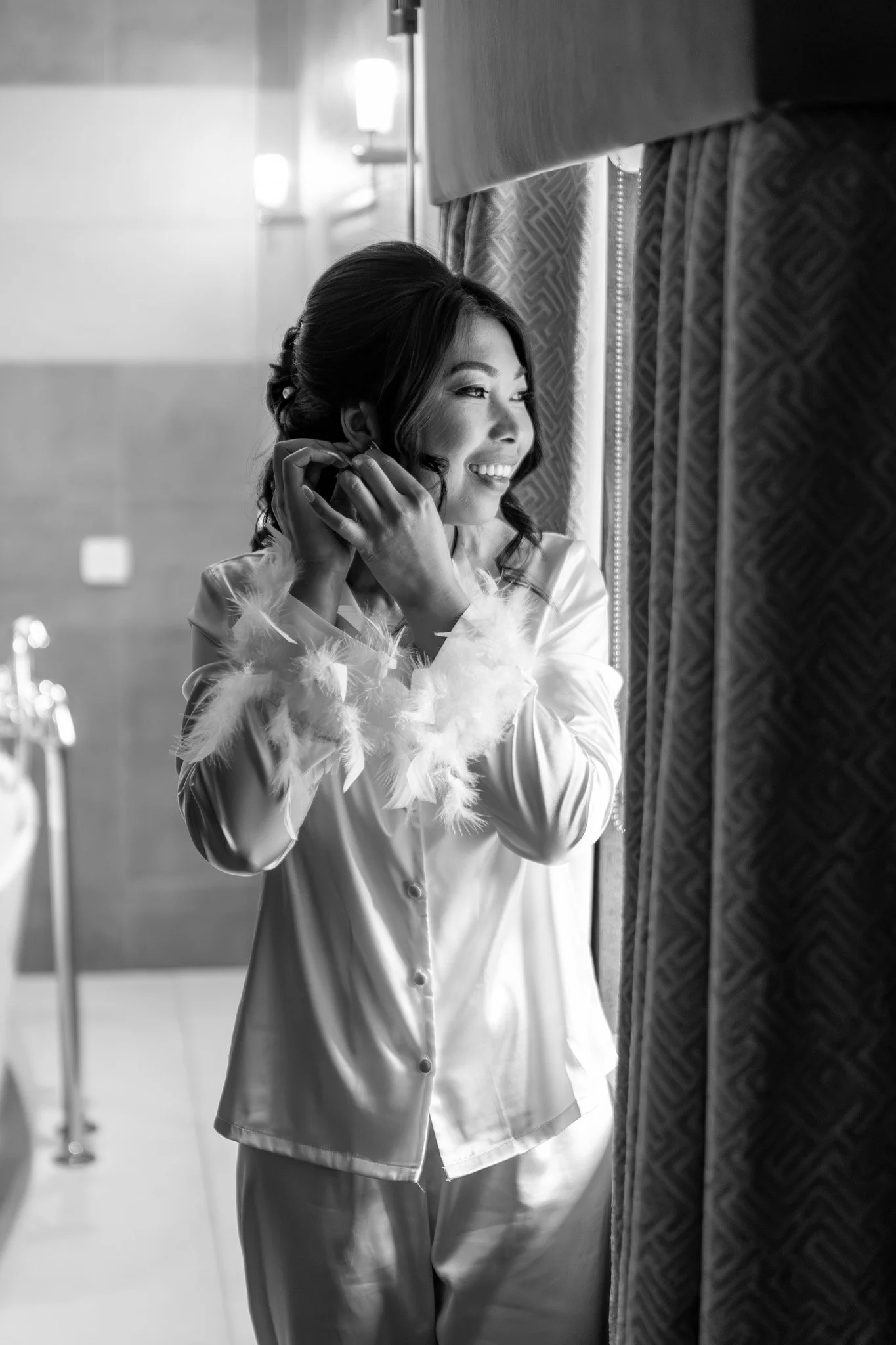 A woman with dark hair in an updo, wearing satin pajamas with feather cuffs, is smiling and putting on earrings while standing by a window with curtains.