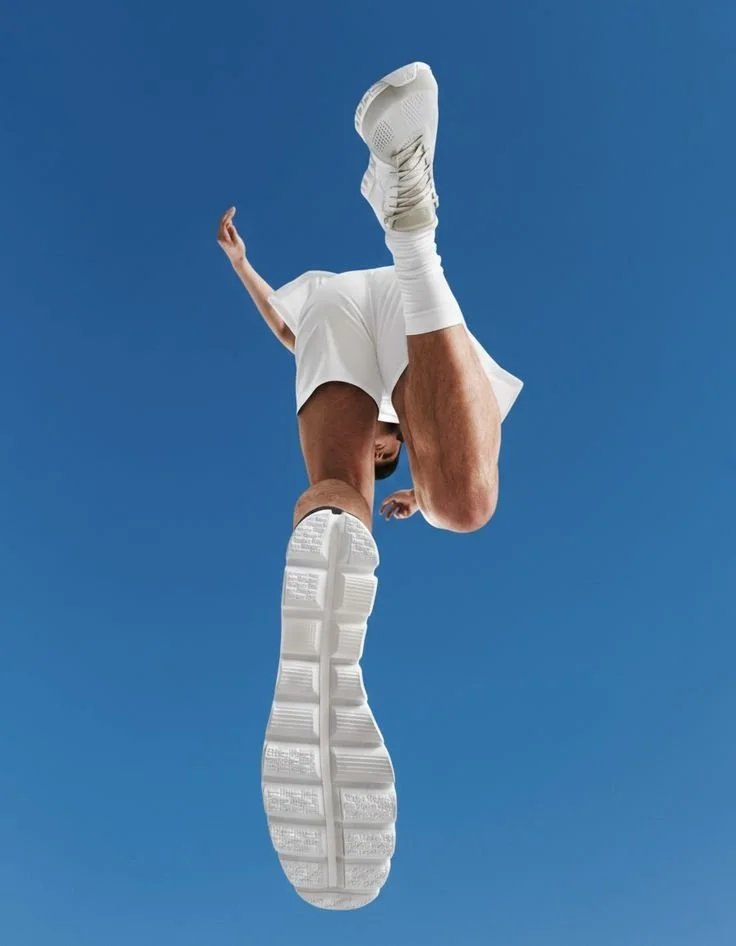 Close-up of a person in white athletic clothing and sneakers, viewed from below against a clear blue sky.