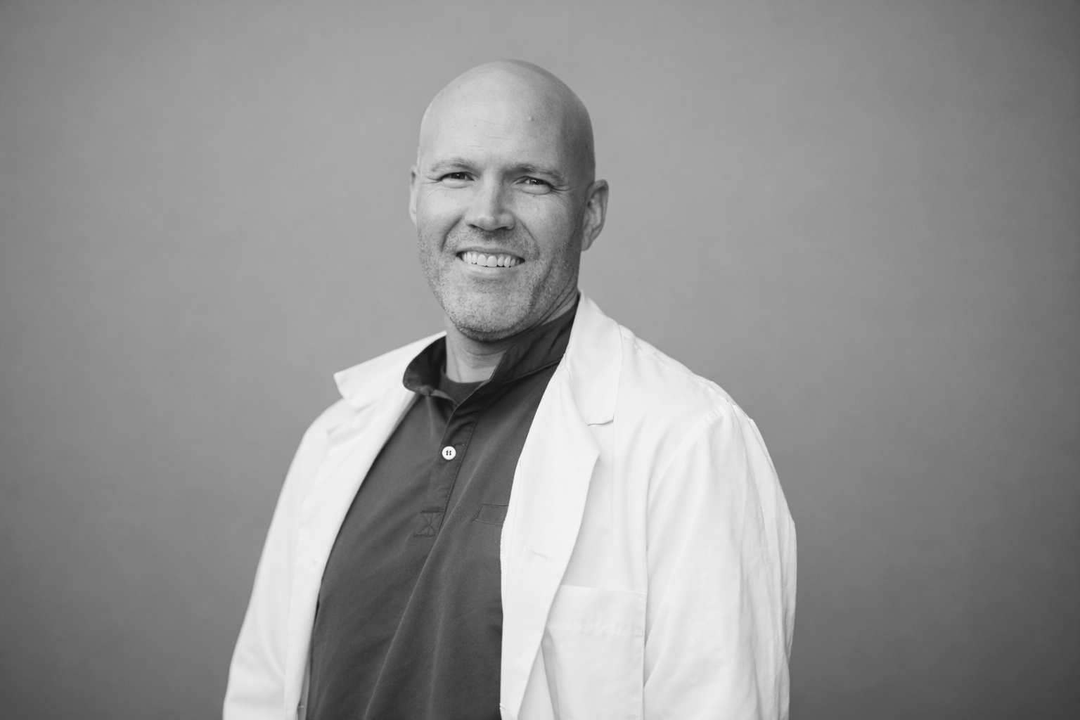A smiling man in a white lab coat standing indoors in front of a blurred background.