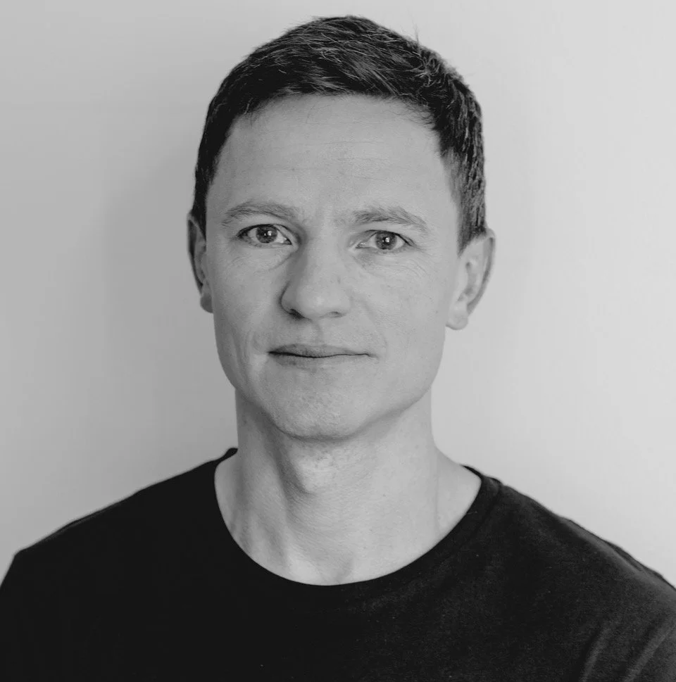 Black-and-white portrait of a man with short dark hair, light skin, wearing a black T-shirt, looking at the camera against a plain background.