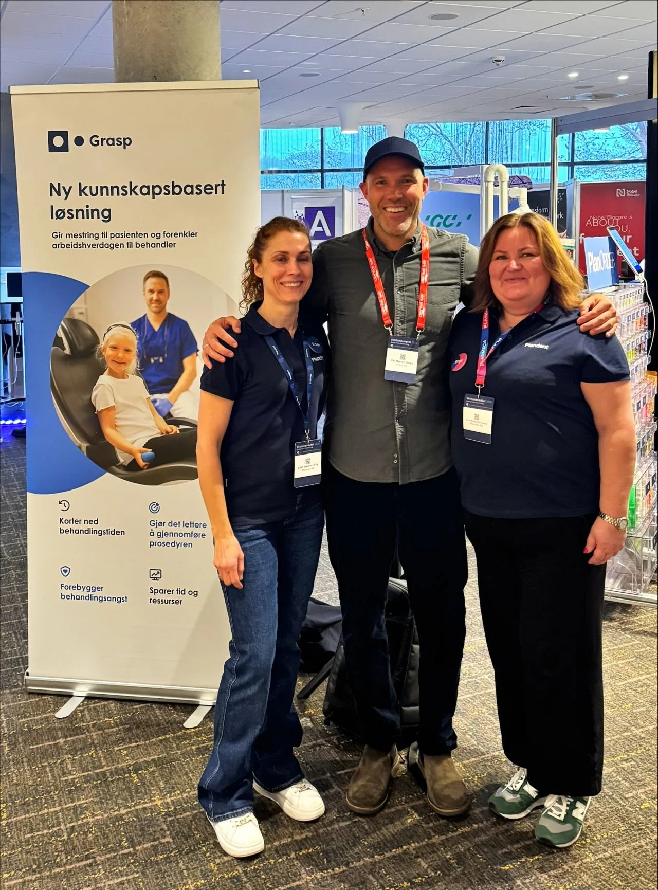 Three people standing together at an event in front of a promotional banner. All are smiling and wearing conference badges. The banner displays a health-related image and text in Norwegian.