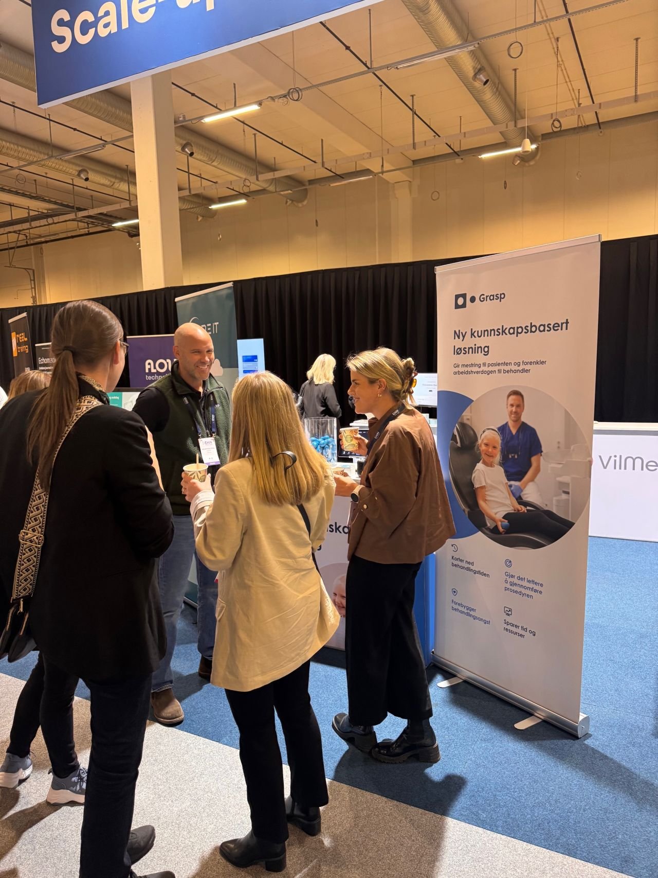 People talking at a booth with a banner that features a smiling child in a dental chair and text in Norwegian promoting a knowledge-based solution for healthcare.