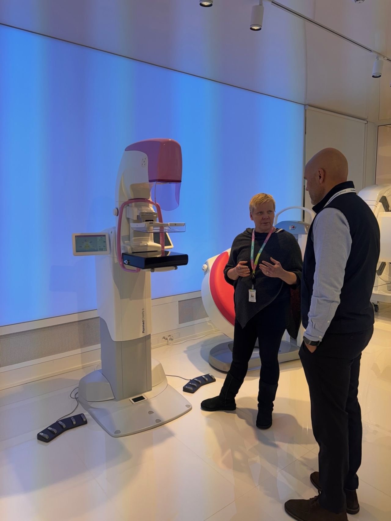 Two people talking next to a large medical imaging machine, possibly an MRI or CT scanner, in a medical facility or showroom.