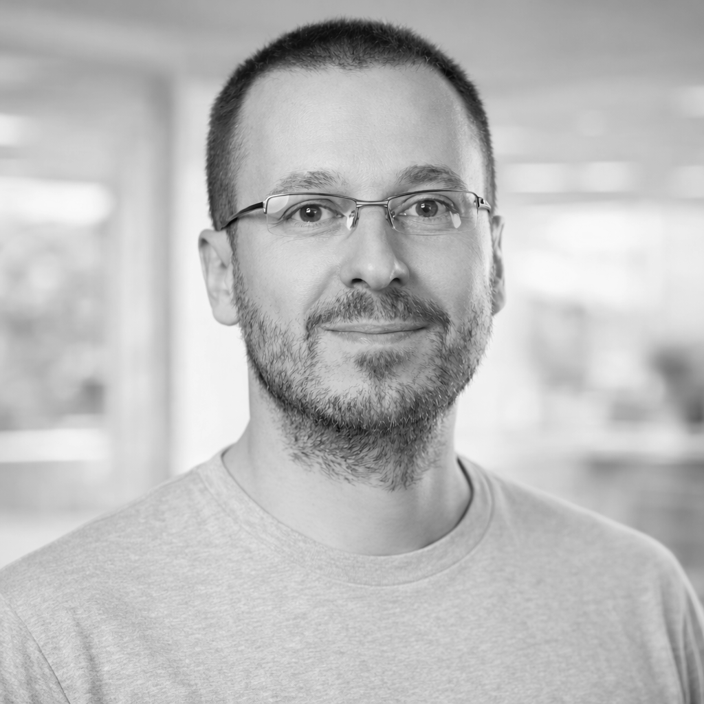 Black and white portrait of a man with short dark hair, glasses, facial hair, wearing a hoodie, looking at the camera.