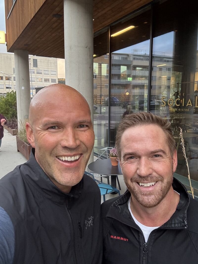 Two smiling men taking a selfie outside a restaurant or café, with large windows and city buildings in the background.