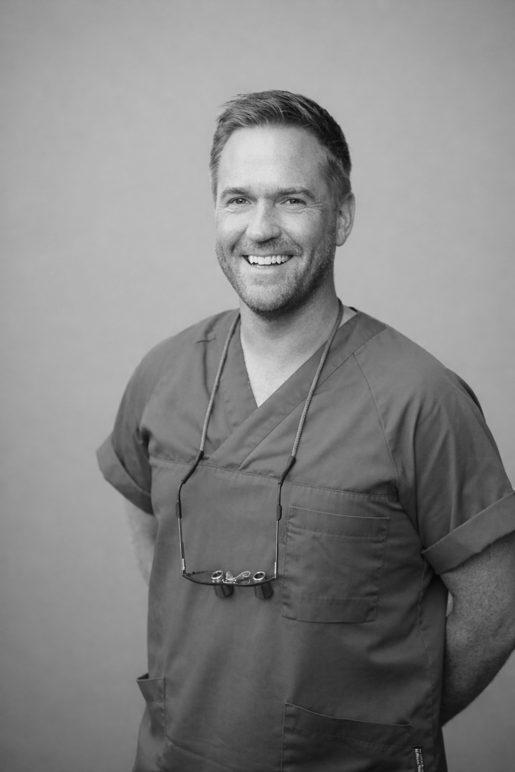 A smiling male healthcare professional in scrubs standing at a doorway, with glasses hanging around his neck.