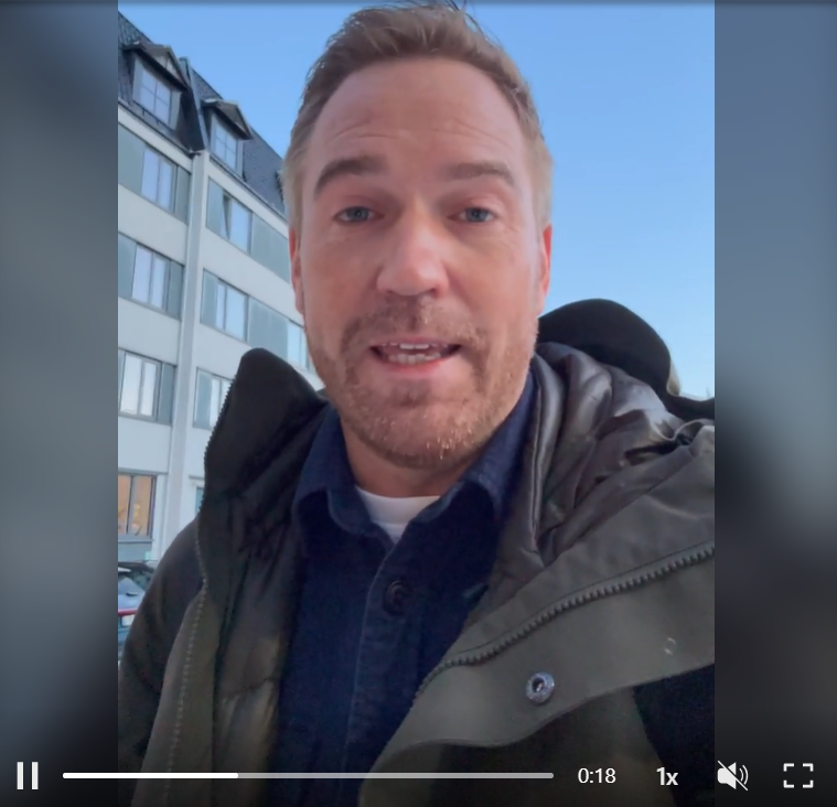 A man with light brown hair and a beard is outside in front of a modern apartment building, wearing a black jacket and blue shirt, speaking to the camera.