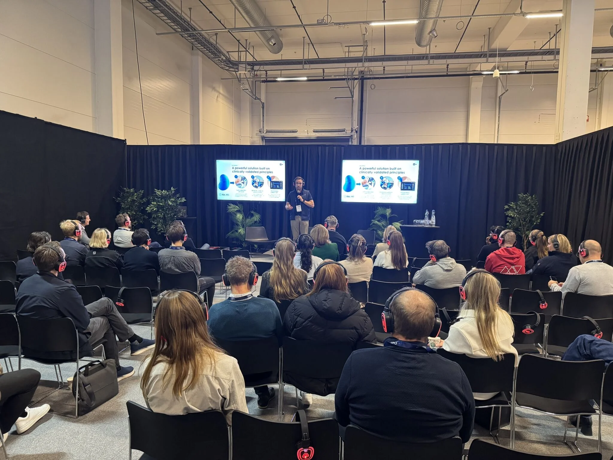 A speaker giving a presentation at a conference with an audience wearing headphones, seated in rows and facing a stage with a large screen behind him, displaying slides.