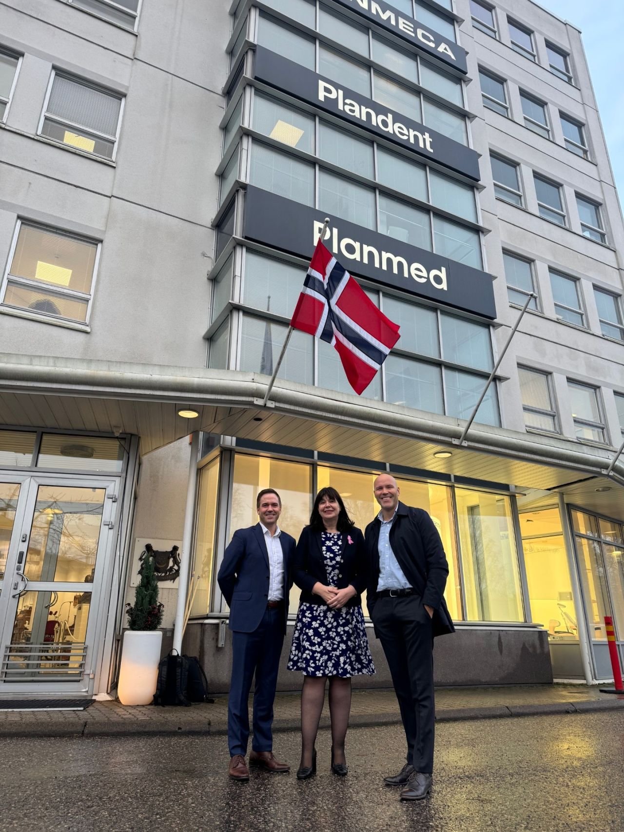 Three people in business attire standing outside a building with large signs that say 'GAMMEGA,' 'Plandent,' and 'Planmed.' A Norway flag is flying above them, and the building has glass windows and a yellow-lit interior.