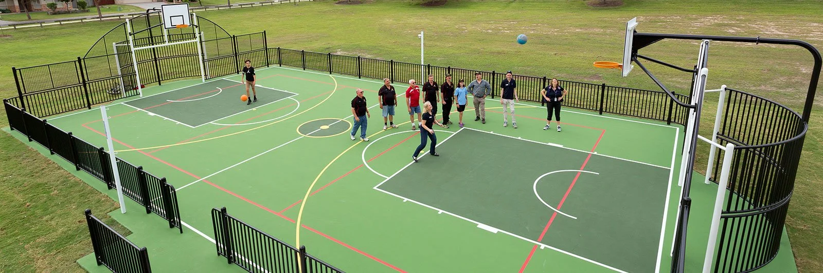 An aerial view of a fenced outdoor multi-sport court with people playing basketball and group of spectators watching, surrounded by green grass.