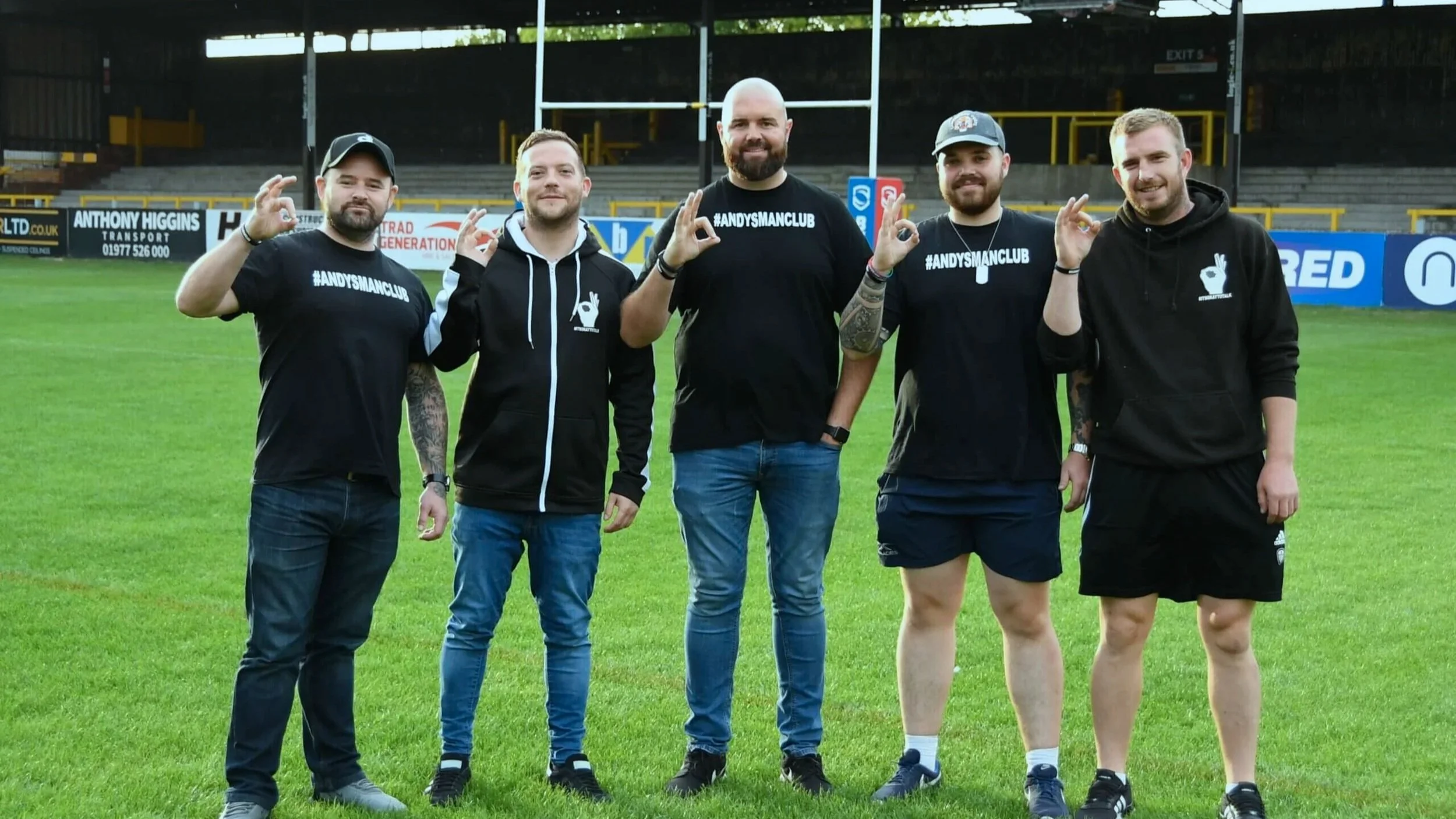 Group of five men standing on a football field, posing for a photo. They are wearing casual and sportswear, with some showing #ANDYSMANCLUB on their shirts. The background shows stadium seating and advertising banners.