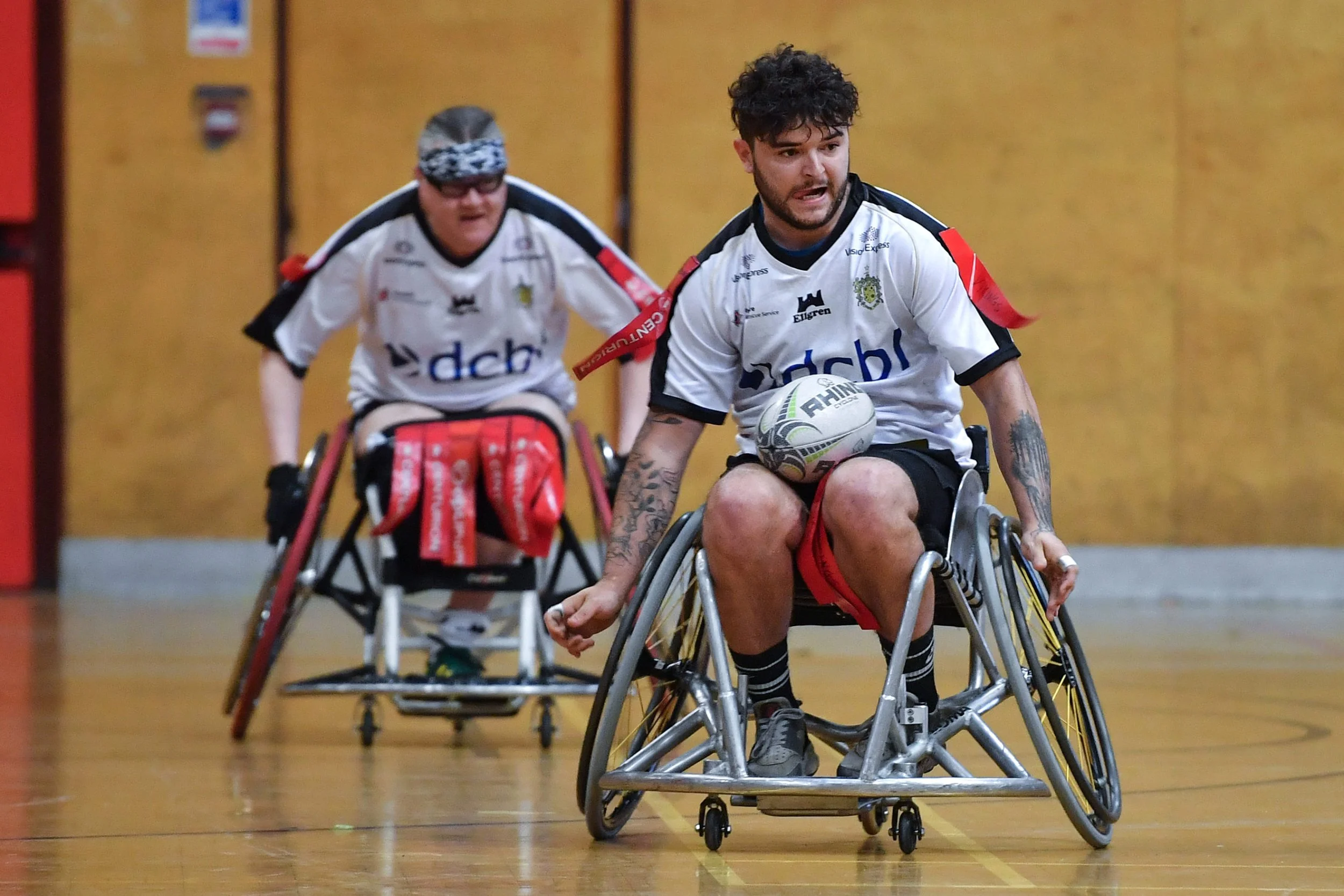 Two male wheelchair rugby players on indoor court, one in the foreground holding a rugby ball, and another in the background, both wearing white jerseys with black accents.