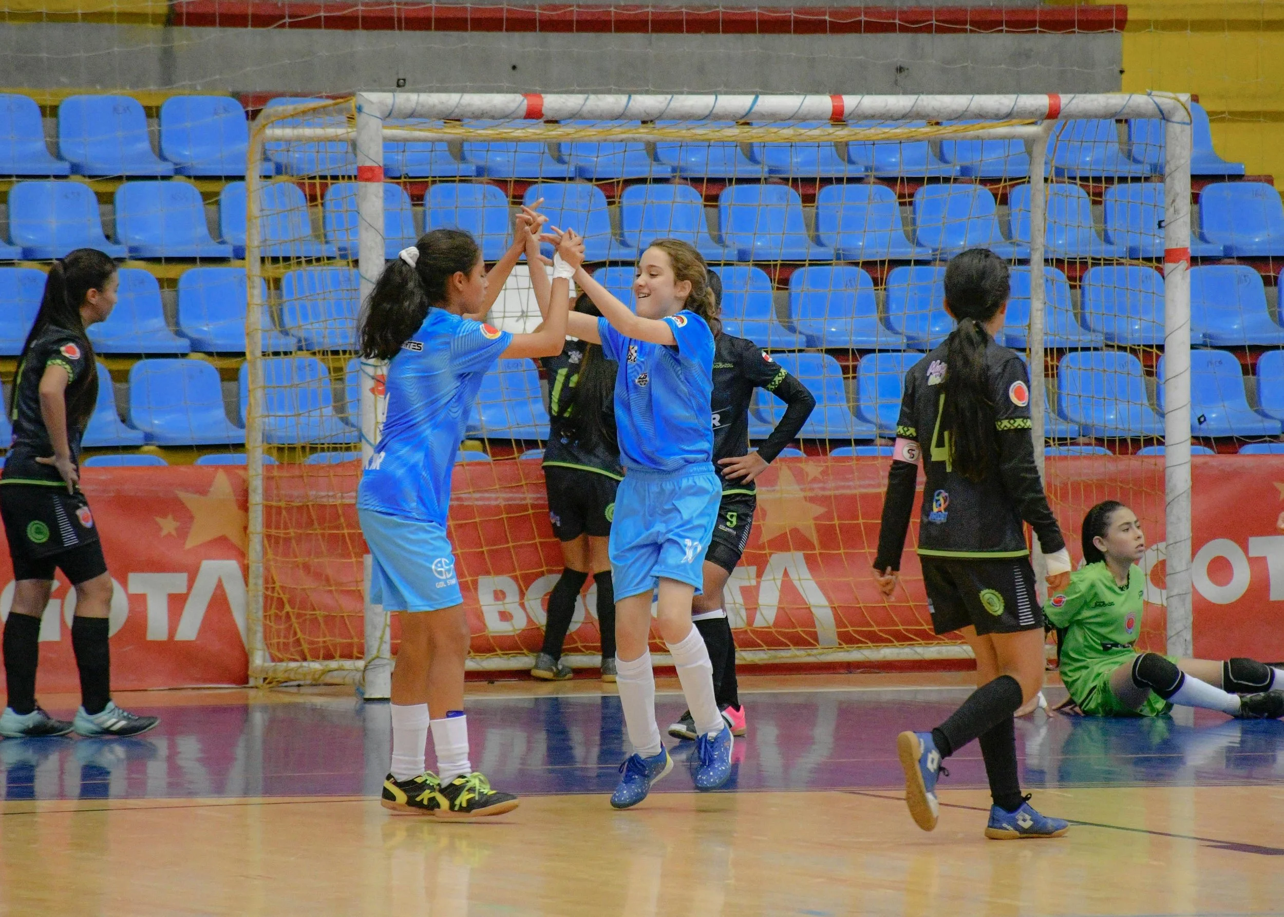 Girls playing indoor soccer, celebrating near the goal, with some girls wearing black jerseys and one in green goalkeeper uniform, inside a sports arena with blue and yellow seats.