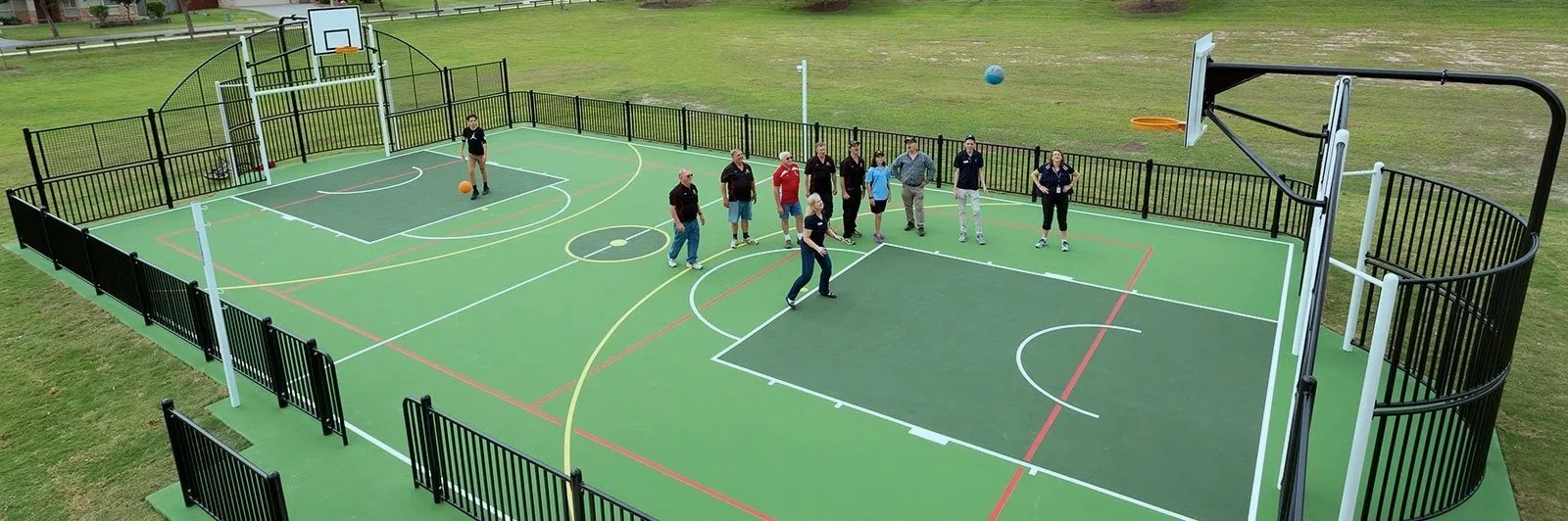 A fenced outdoor multi-sport court with people gathered around. The court has basketball hoops, painted lines for multiple sports, and several individuals, some of whom are holding or near soccer balls, suggesting a recreational or training activity.
