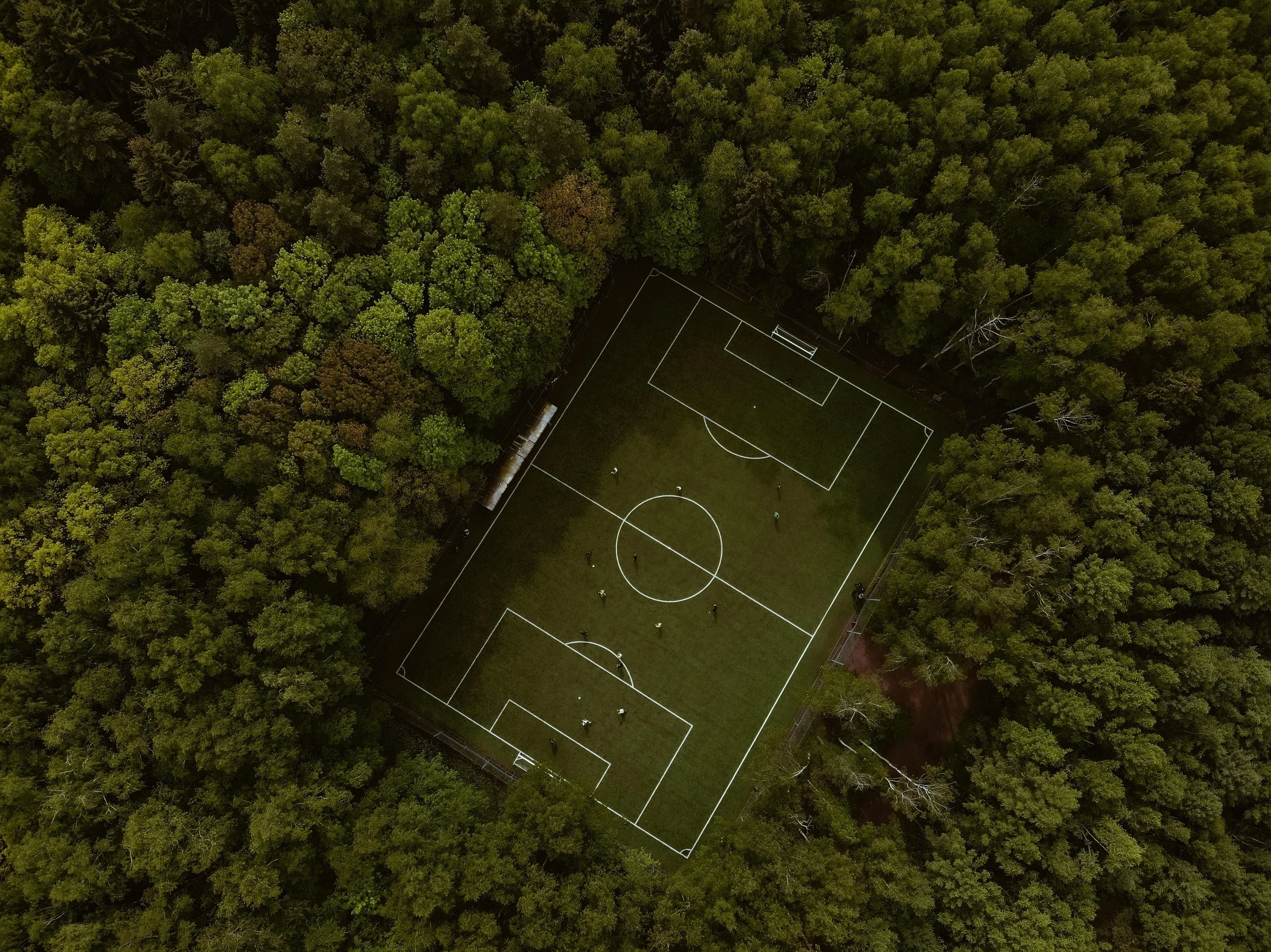 An aerial view of a small soccer field surrounded by dense green trees, with a few people playing on the field.