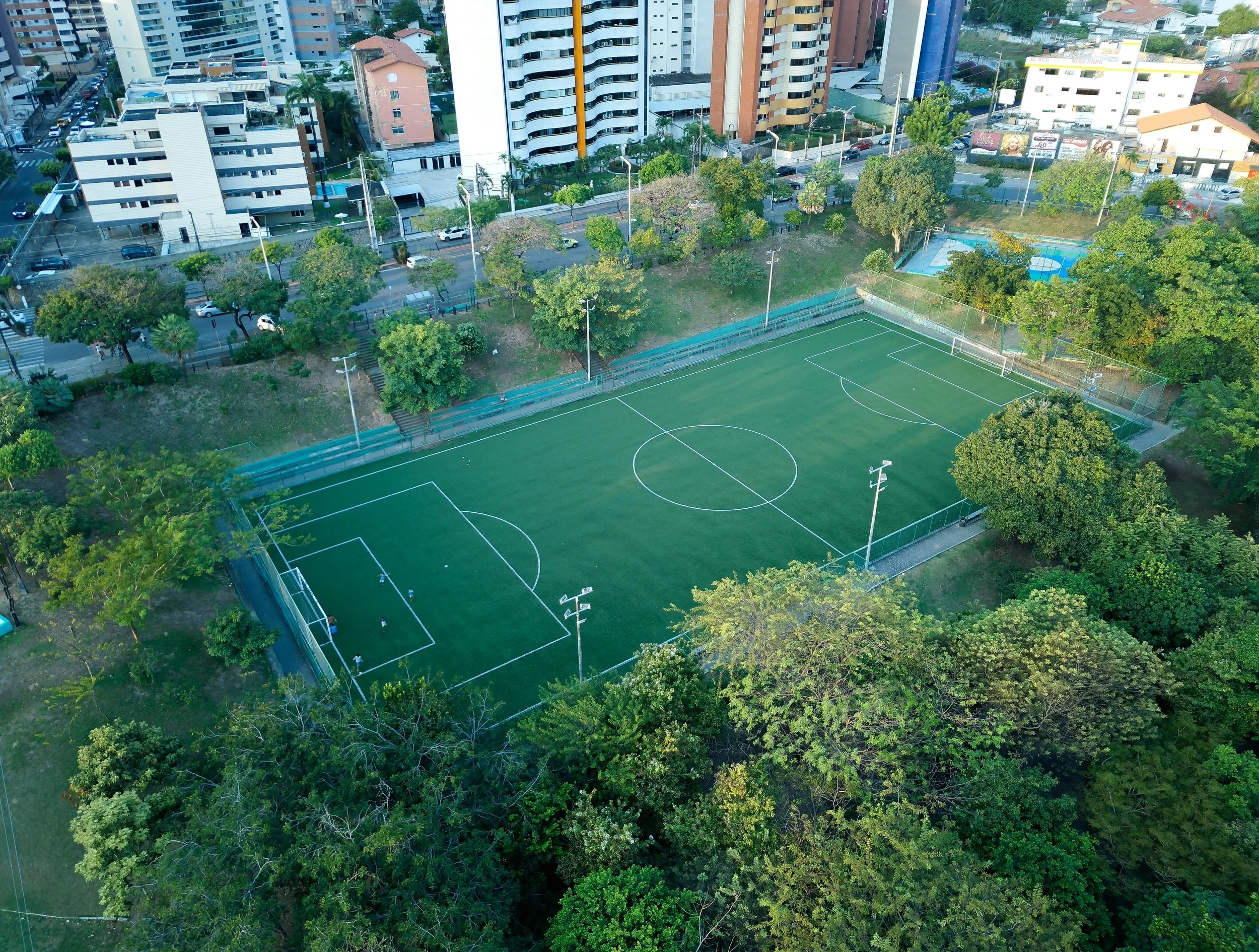 An aerial view of a green soccer field surrounded by trees, with residential and commercial buildings in the background.