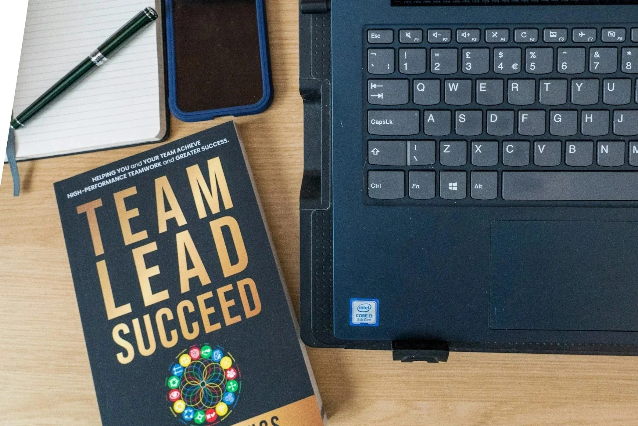 Työpöydällä on kannettava tietokone, älypuhelin, muistiinpanovihko ja pyyhekynä, sekä teosta ”Team Lead Succeed”