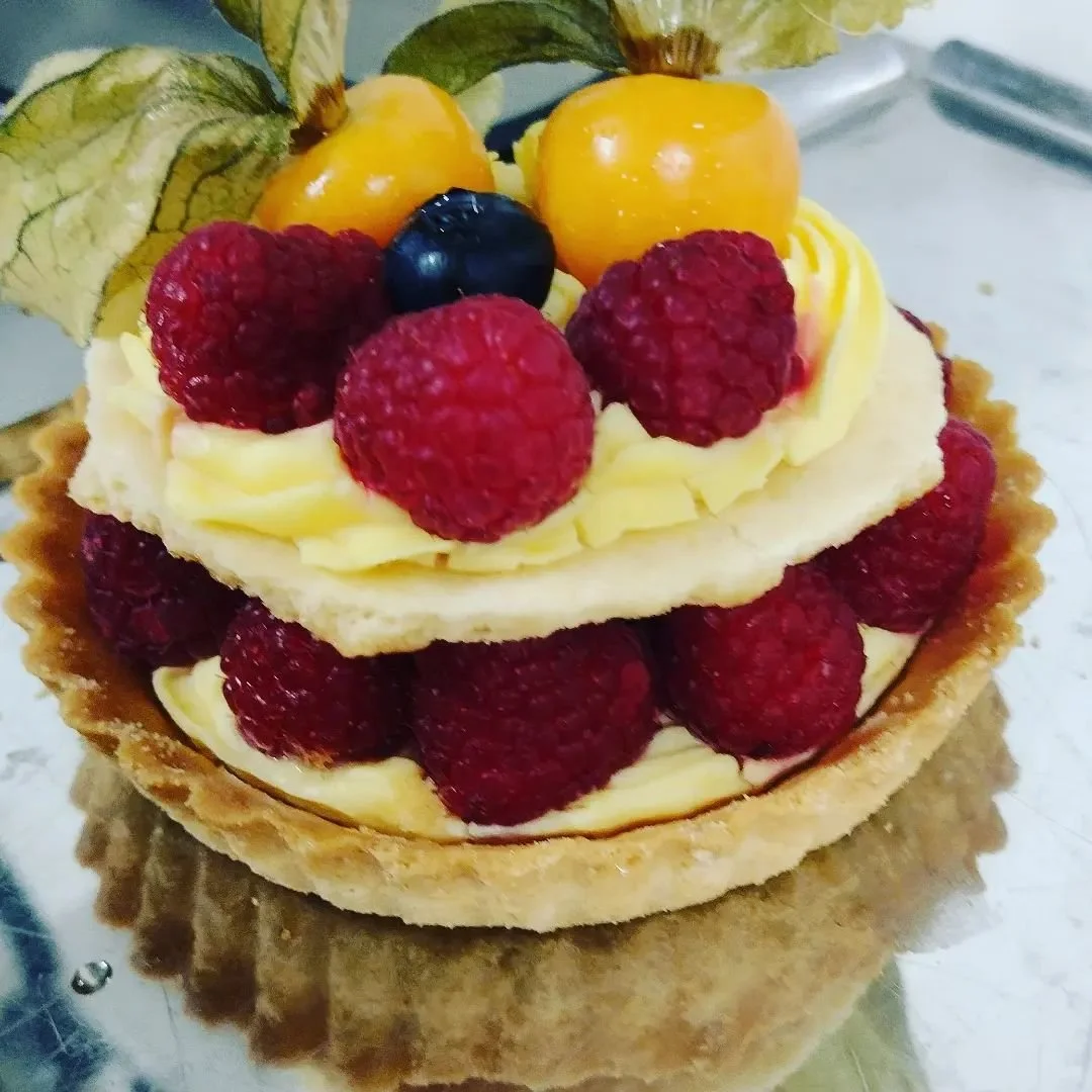 A fruit tart with a pastry crust, topped with layers of raspberries, blueberries, yellow gooseberries, and cream, garnished with a physalis on top.