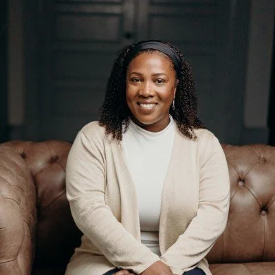 Smiling woman sitting on a brown leather couch in a cozy, dark-toned room.
