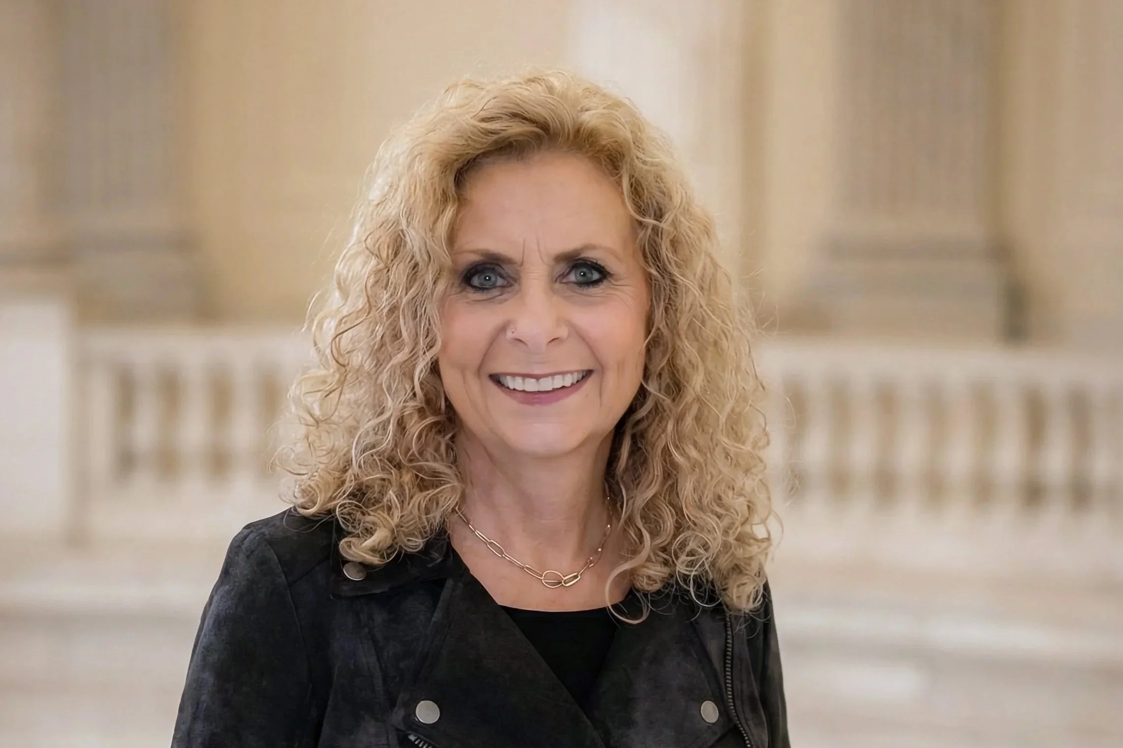 A woman with curly blonde hair, blue eyes, and a bright smile, wearing a black jacket and a gold necklace, standing in a grand building with tall columns in the background.