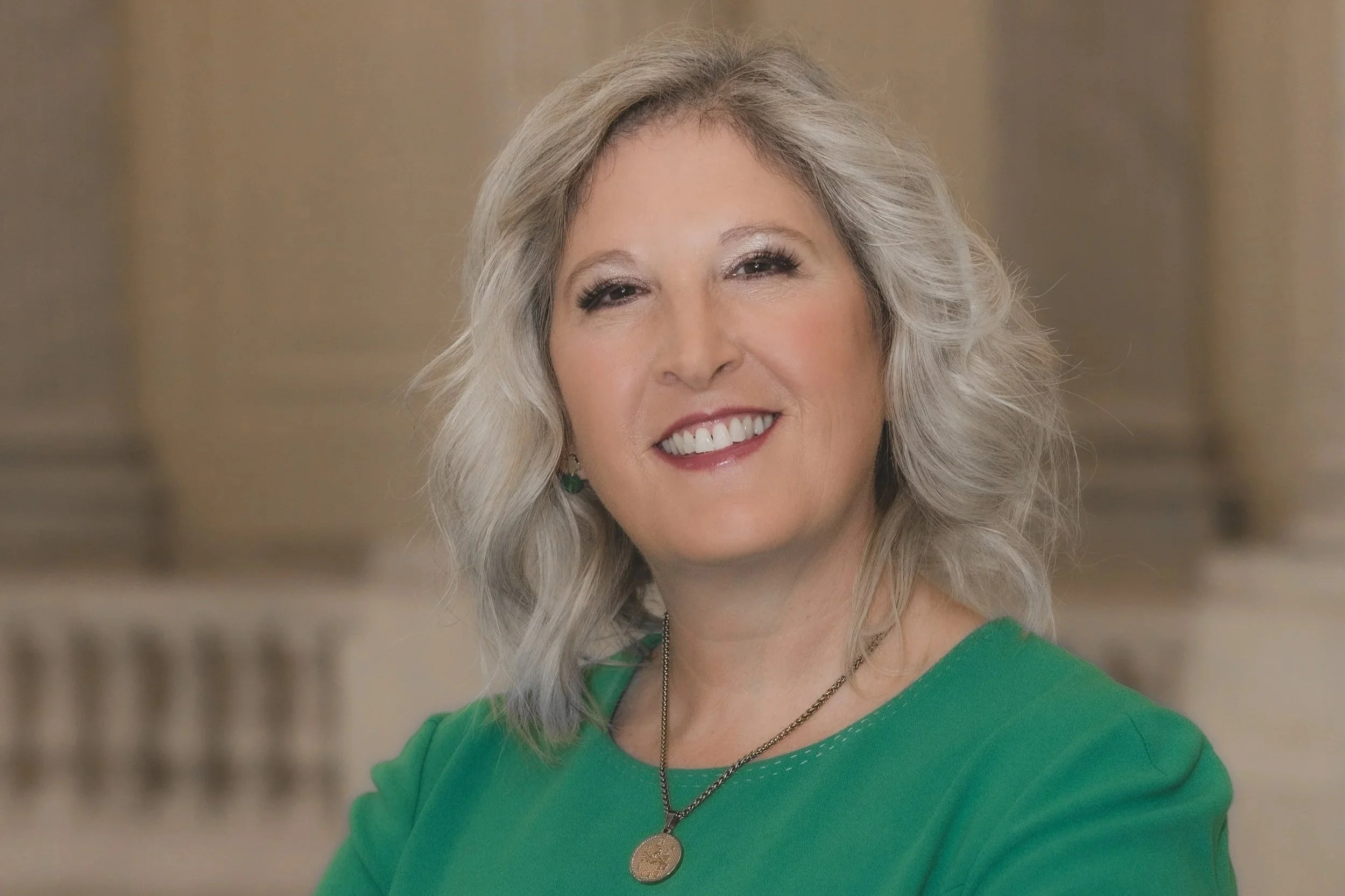 Portrait of a smiling woman with wavy gray hair wearing a green top and a gold necklace with a round pendant, indoors with classical architecture in the background.