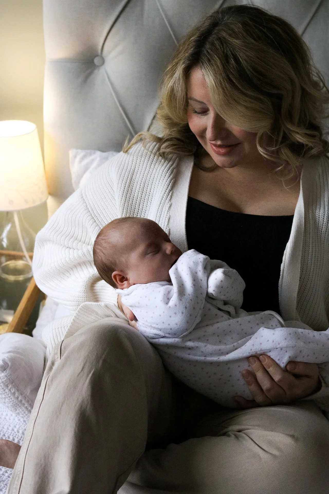 A woman holding a sleeping baby in her lap on a bed, with a lamp on a nightstand in the background.