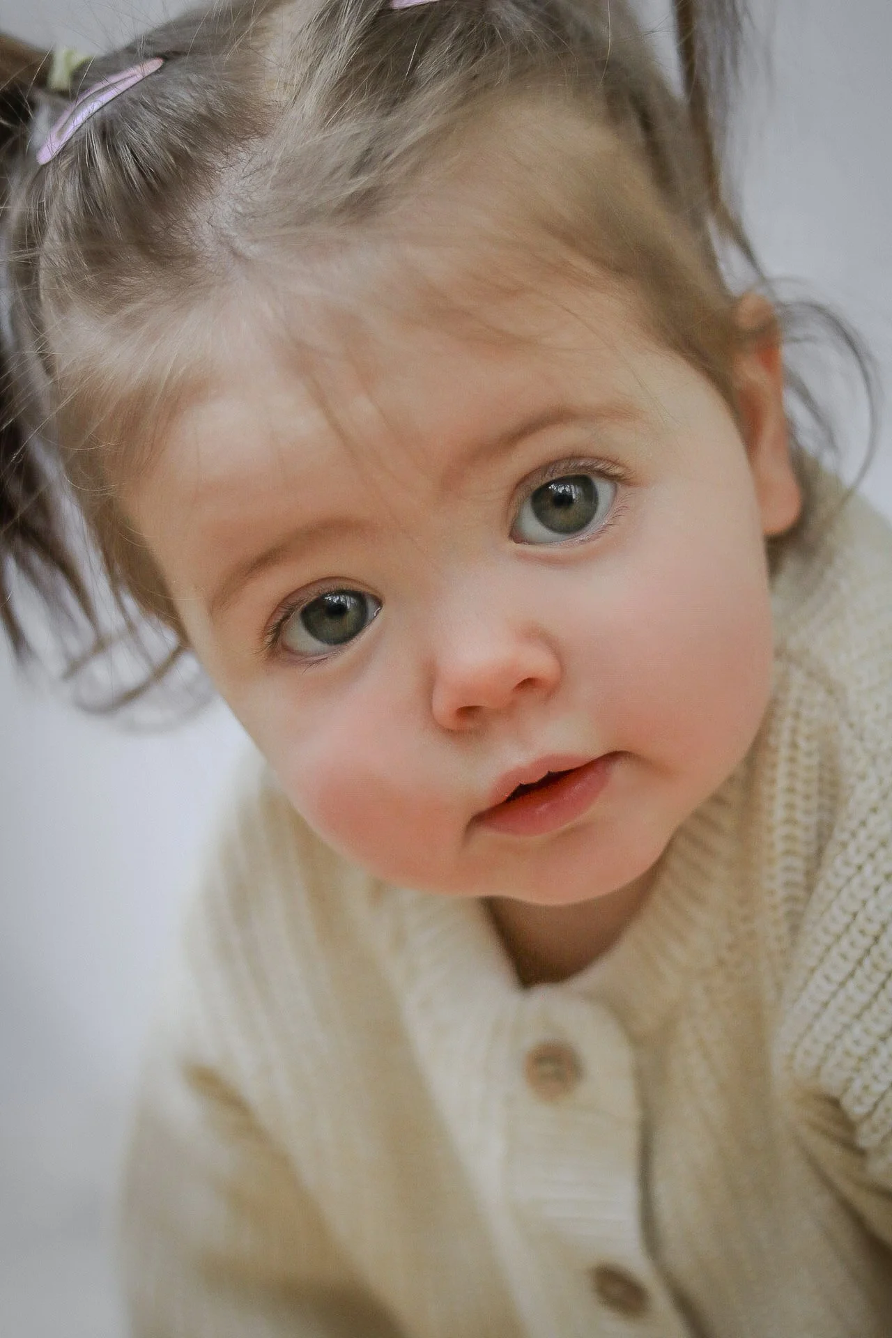 Close-up of a young child with large gray eyes, light brown hair pulled back with a purple hair clip, and wearing a cream-colored knitted cardigan.
