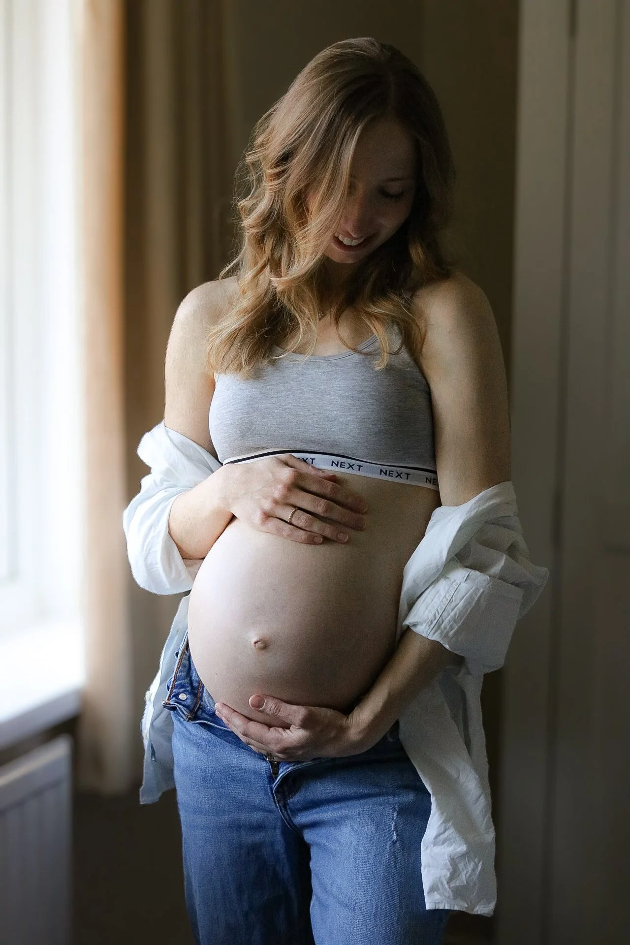 A pregnant woman smiling and touching her belly as she looks down, standing indoors near a window with beige curtains.