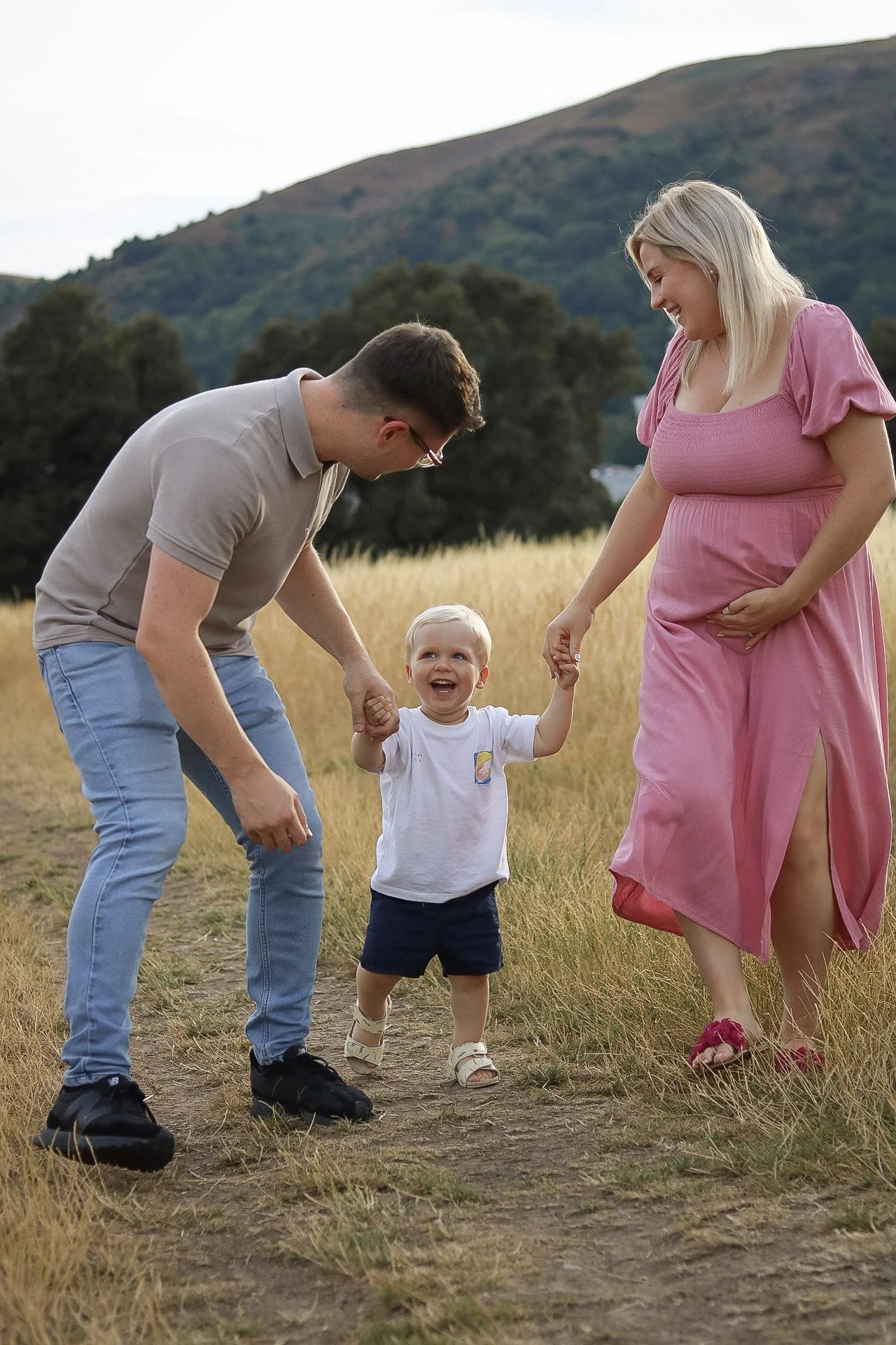 A happy family of three holding hands and walking through a grassy field with mountains in the background, featuring a smiling toddler between the parents.