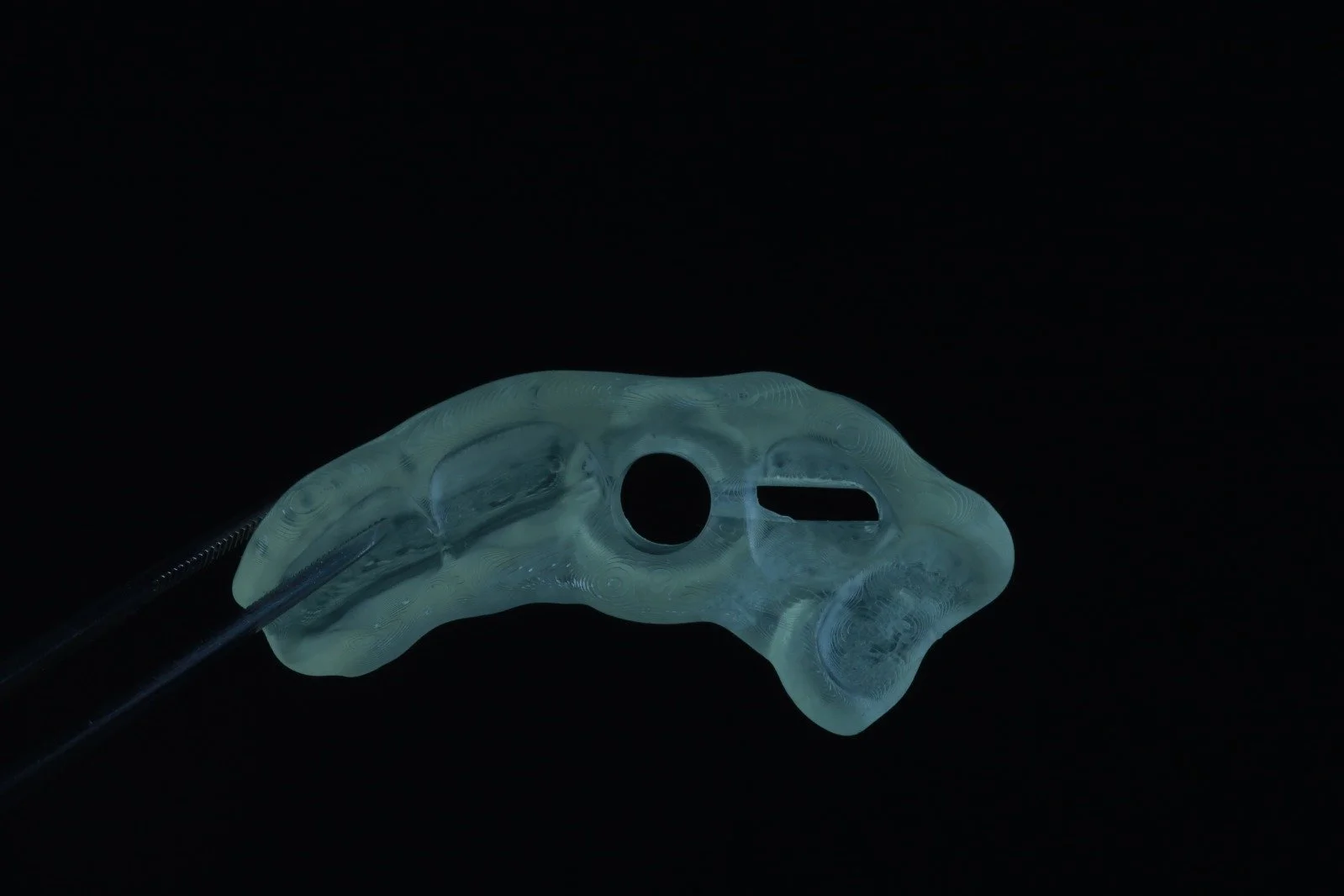 A translucent, abstract, irregularly shaped object with holes, held by a thin rod against a black background.