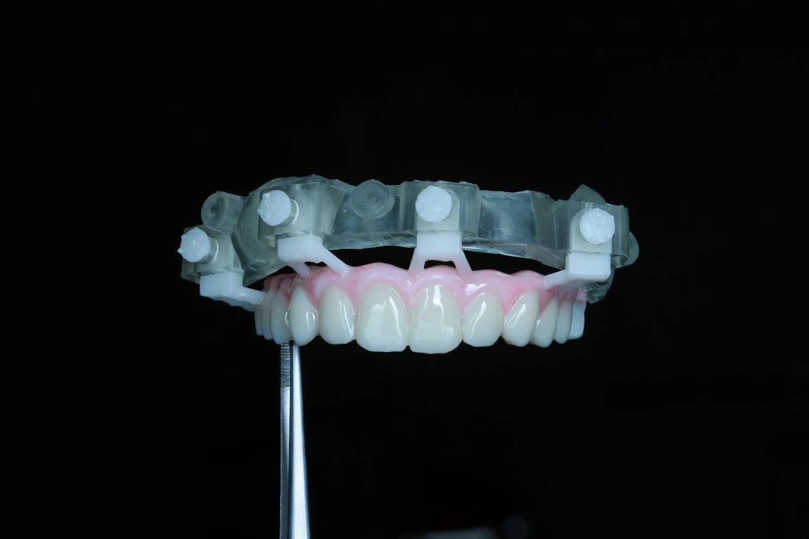 Dental model with upper teeth, pink gums, and clear orthodontic brackets, held by a metal instrument against a black background.